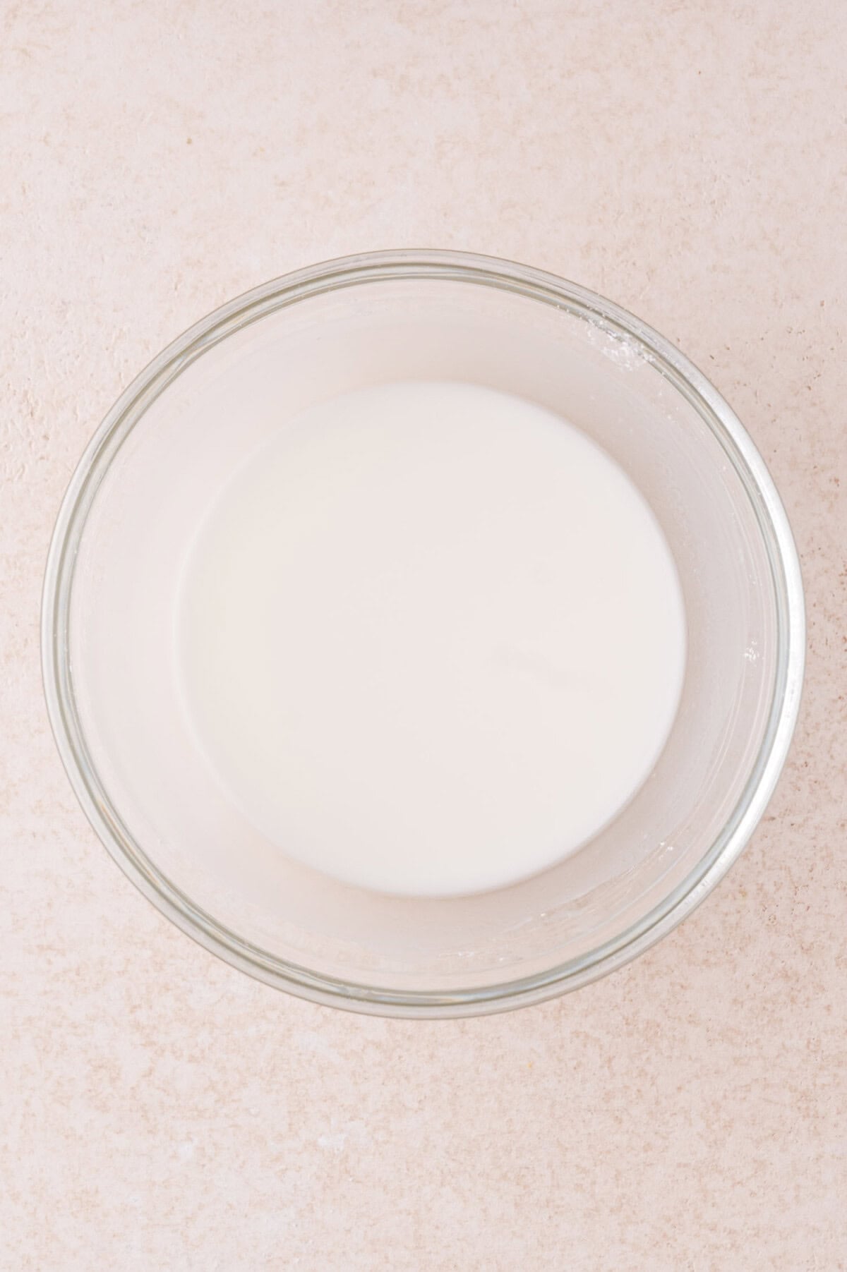 A clear glass bowl filled with a white cornstarch slurry, sits on a light-colored textured surface.