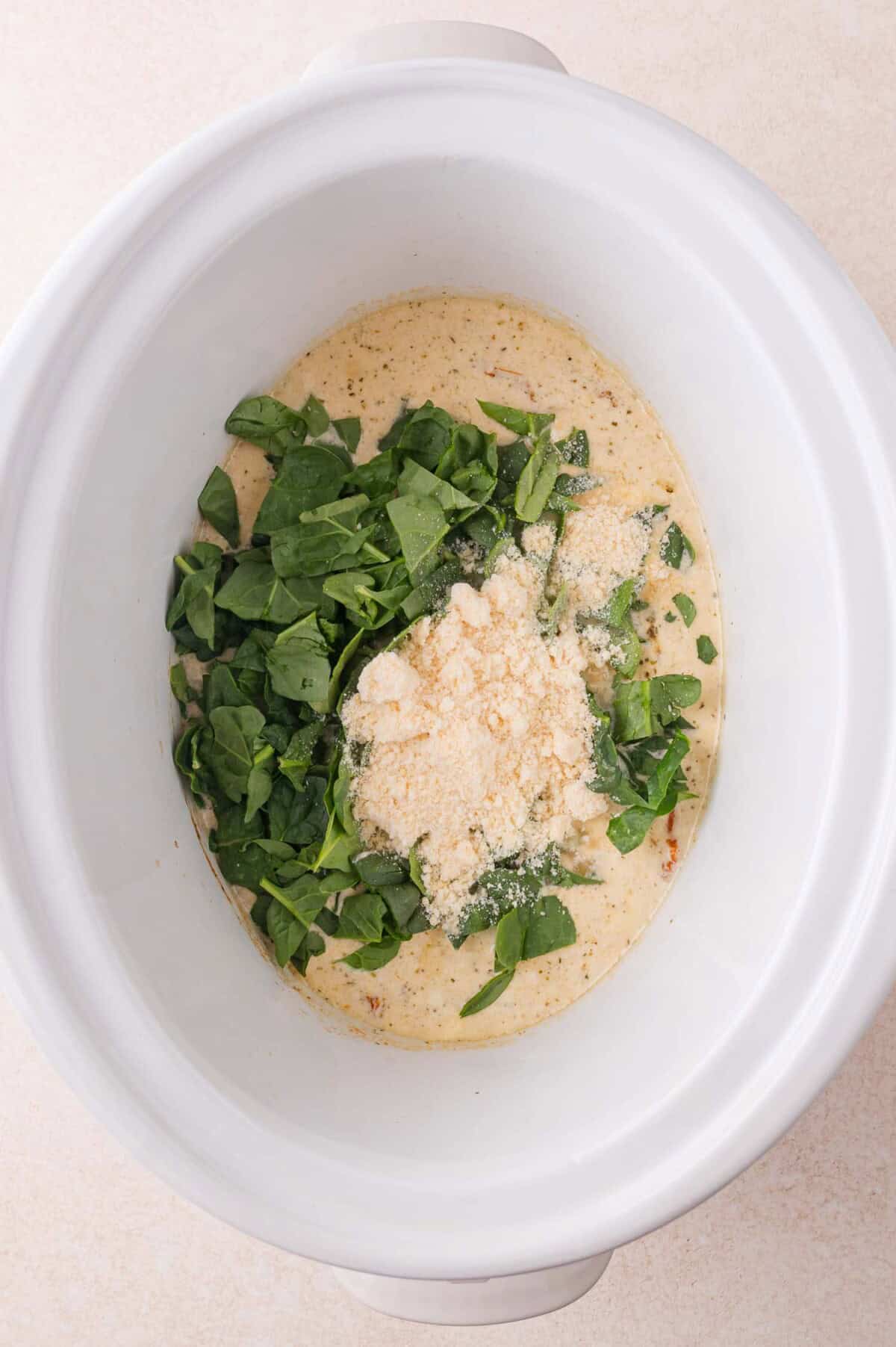 White slow cooker containing a creamy mixture topped with chopped spinach and grated cheese, ready to be mixed or cooked.