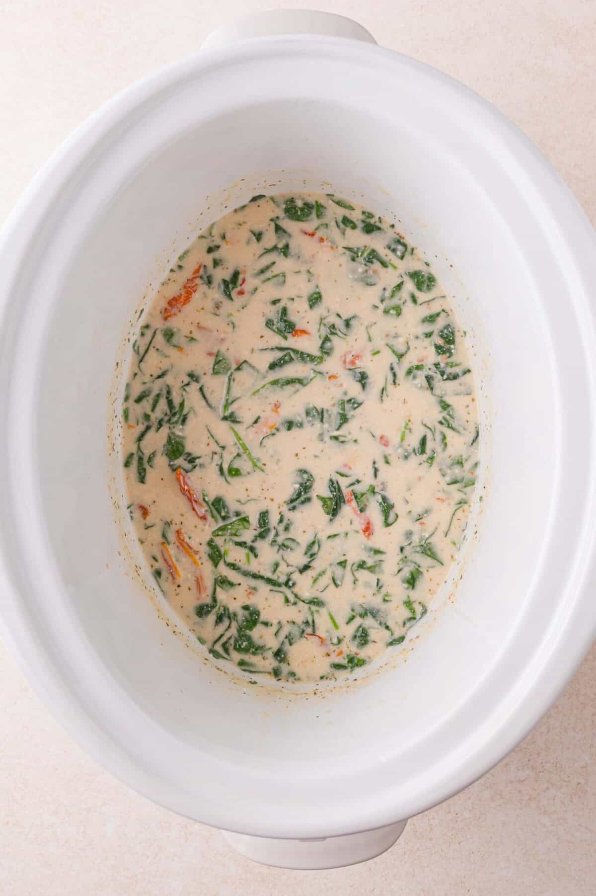 A creamy sauce with chopped spinach and sun dried tomatoes sits in a white slow cooker.