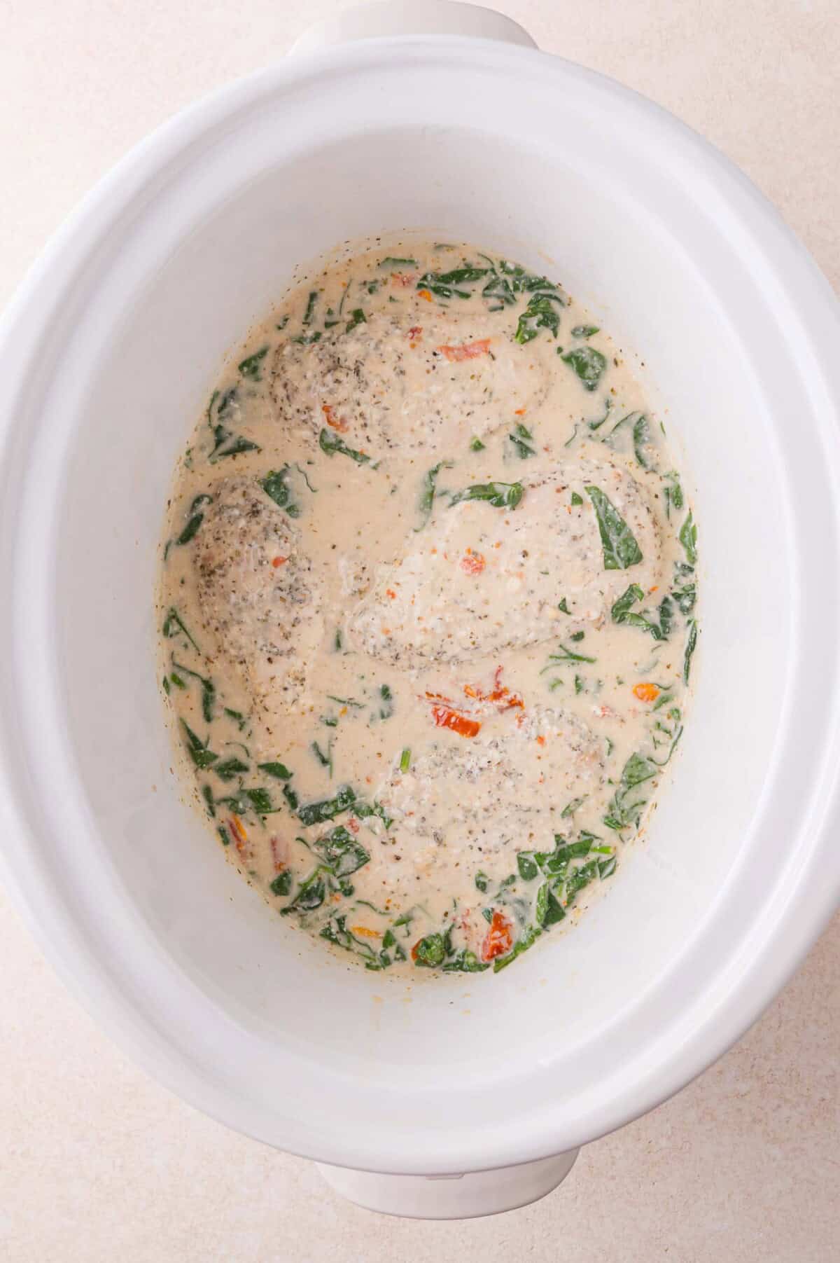 Chicken breasts in a creamy Tuscan sauce with spinach and sun-dried tomatoes in a white slow cooker.