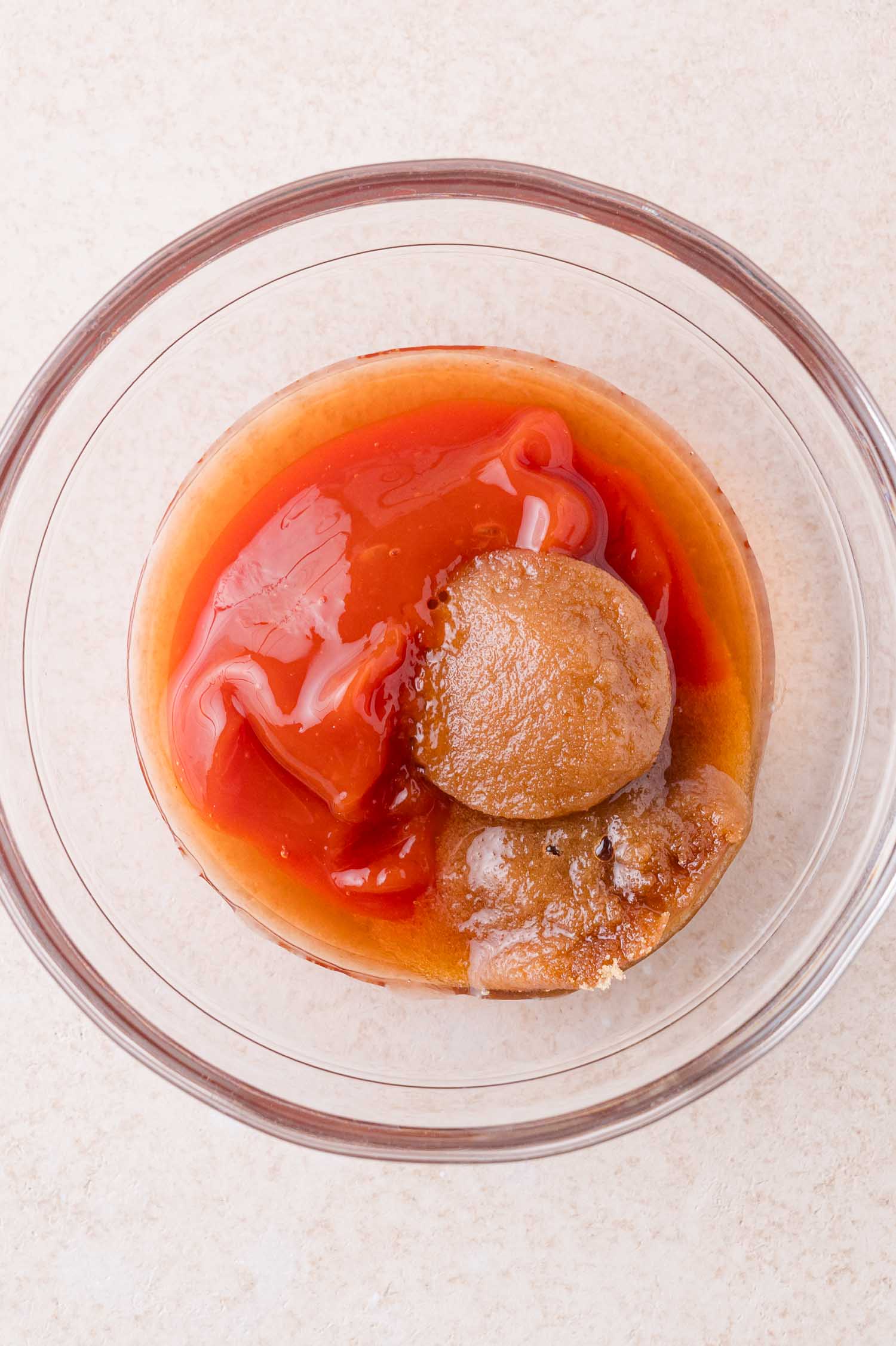 A glass bowl containing brown sugar, ketchup, and other liquid ingredients on a light-colored surface.