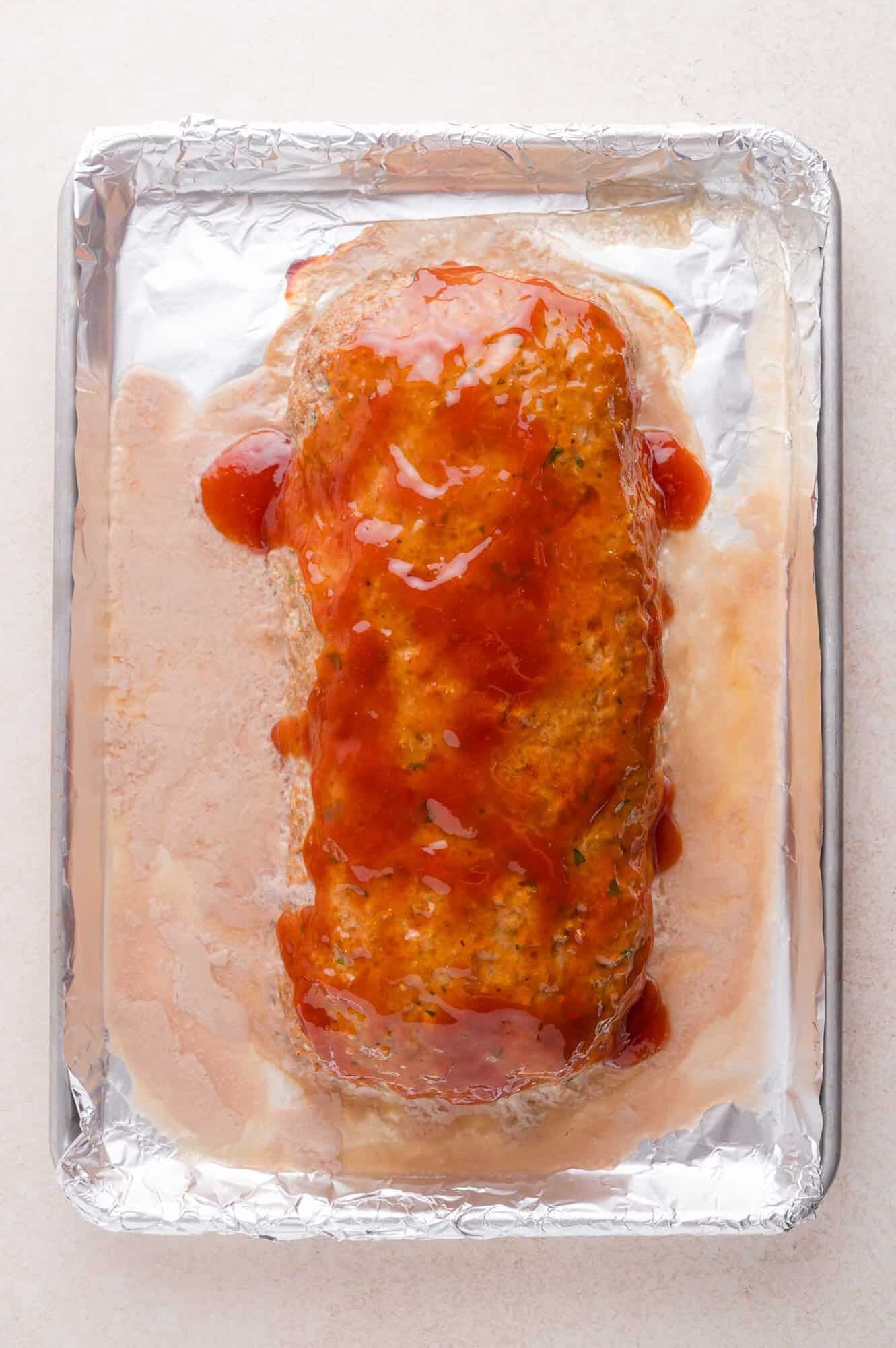 A baked meatloaf covered in ketchup glaze sits on a foil-lined baking sheet with juices pooling around it.
