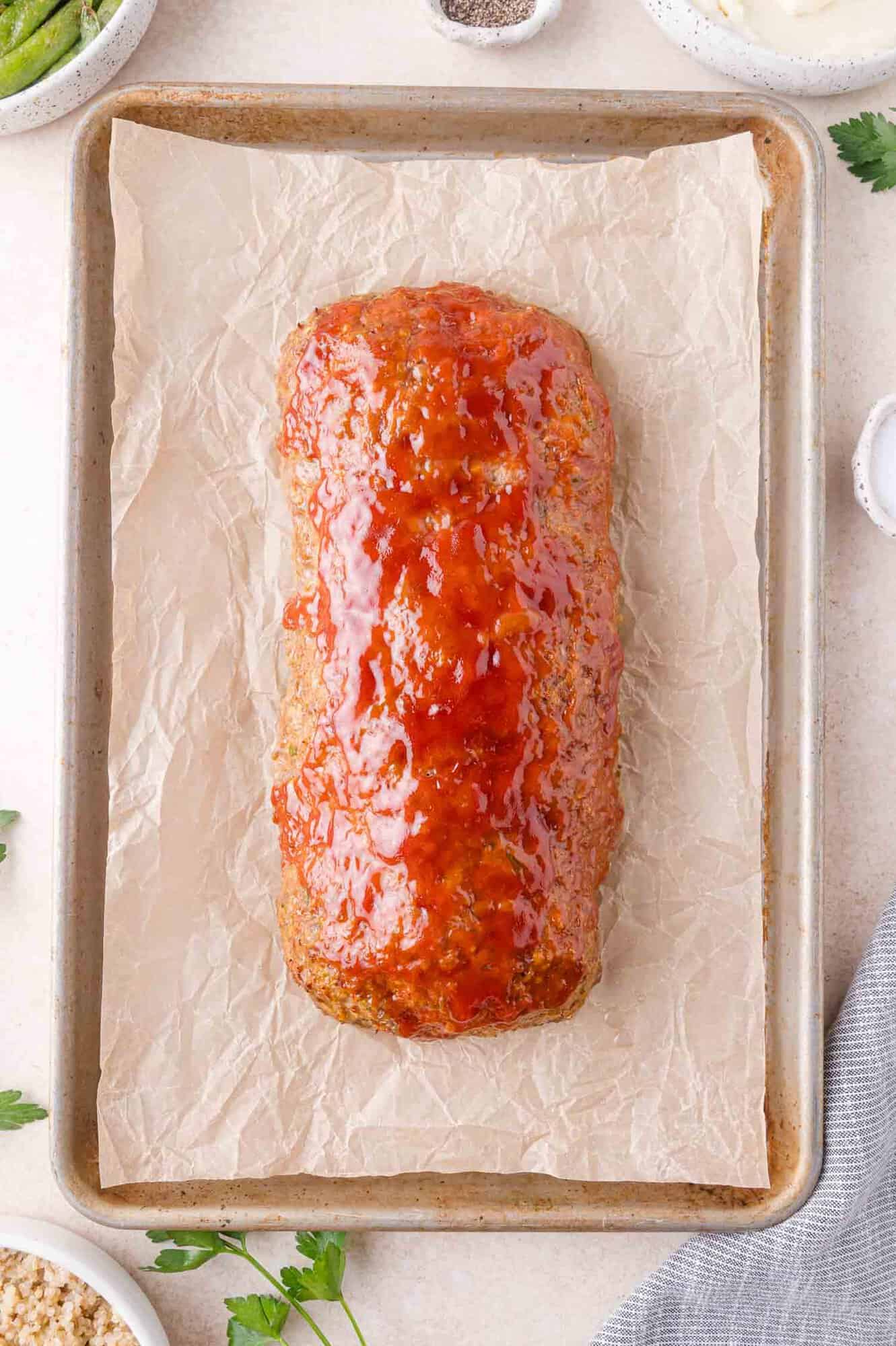 A glazed meatloaf sits on a parchment-lined baking sheet, surrounded by parsley and small bowls of ingredients.