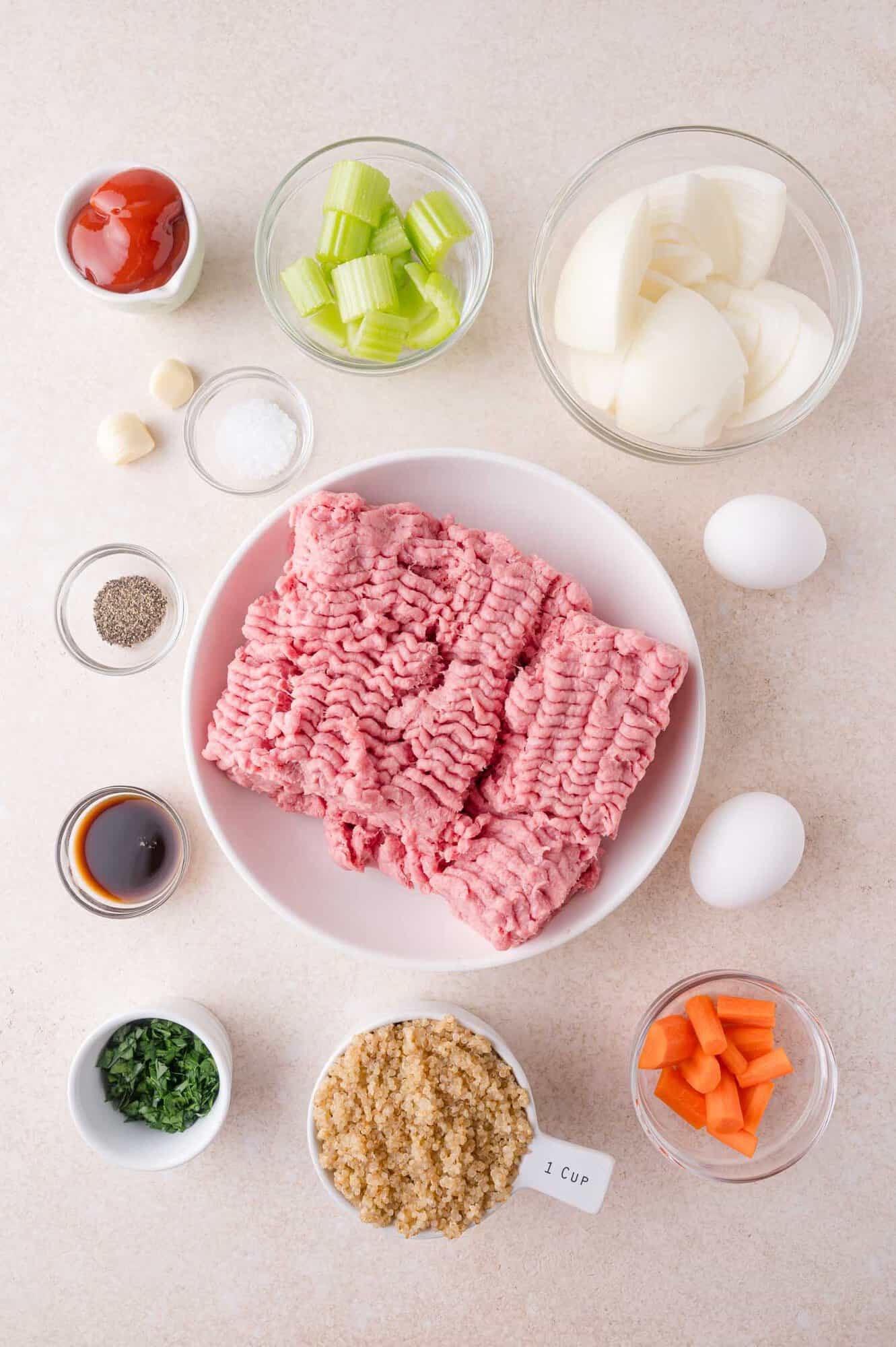 A plate of raw ground turkey is surrounded by bowls with diced onion, celery, carrot, bread crumbs, parsley, ketchup, Worcestershire, salt, pepper, garlic, and two eggs on a light surface.