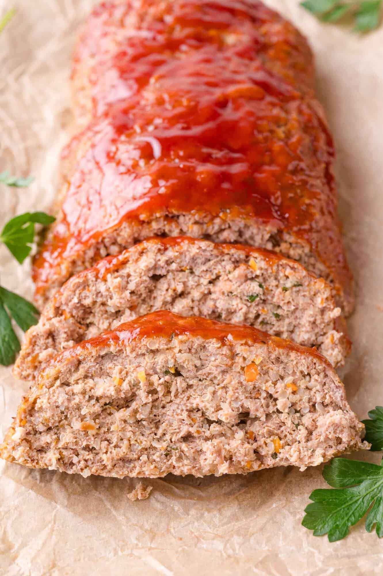 Sliced meatloaf with a shiny tomato glaze on top, served on parchment paper with parsley garnishes.
