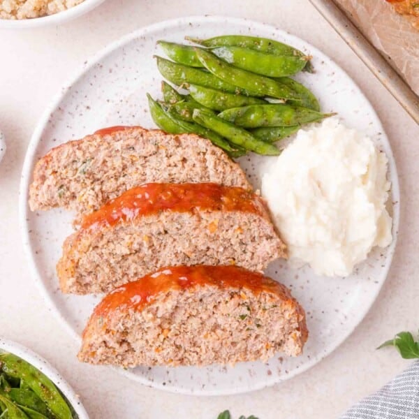 Three slices of turkey quinoa meatloaf, mashed potatoes, and seasoned snap peas arranged on a white speckled plate.