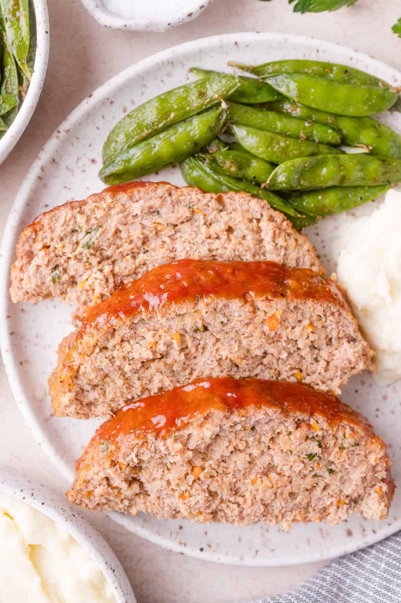 Three slices of glazed meatloaf served with mashed potatoes and green beans on a white plate.