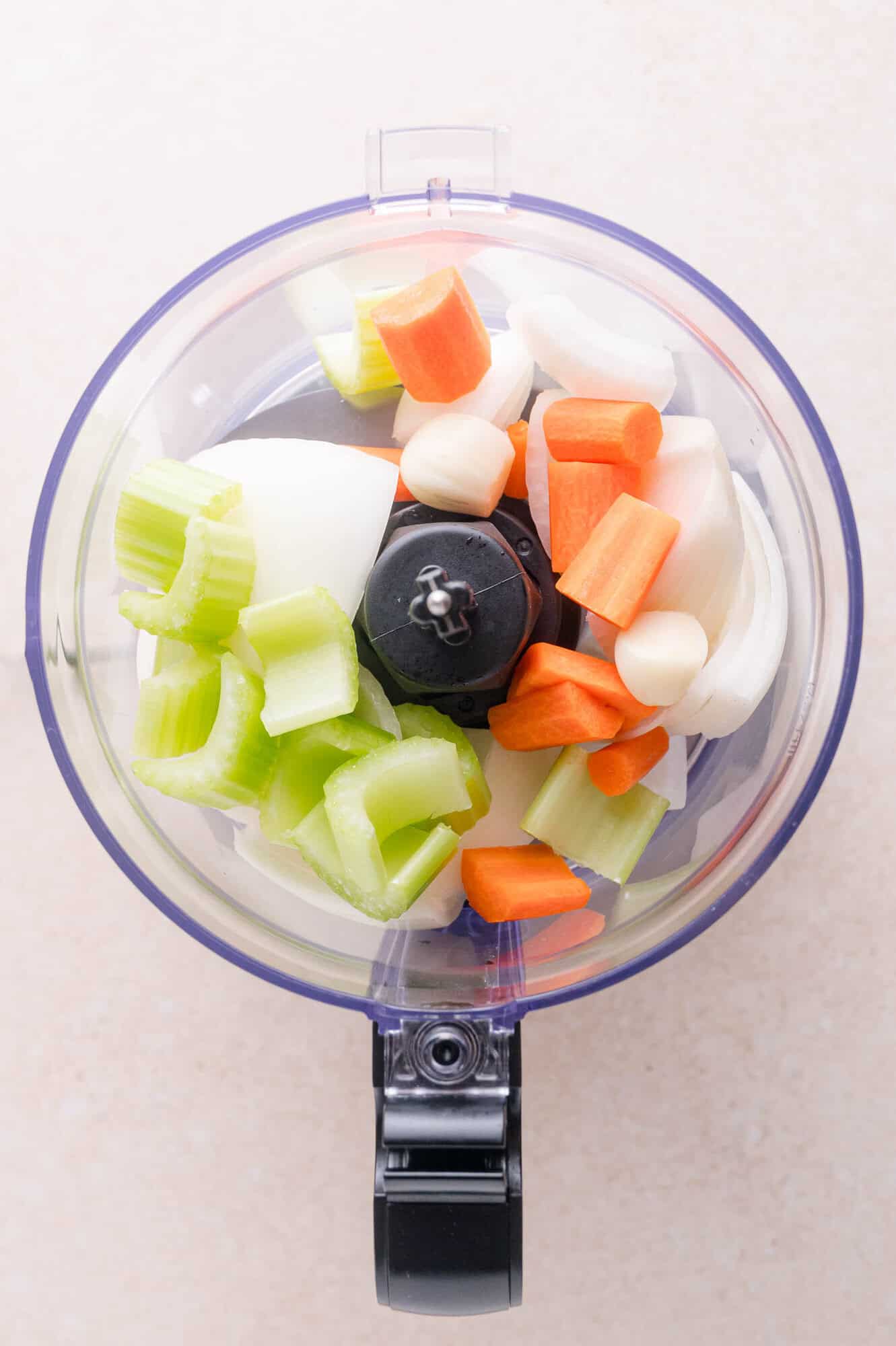 Chopped celery, onion, carrots, and garlic in a food processor bowl, viewed from above on a light-colored surface.