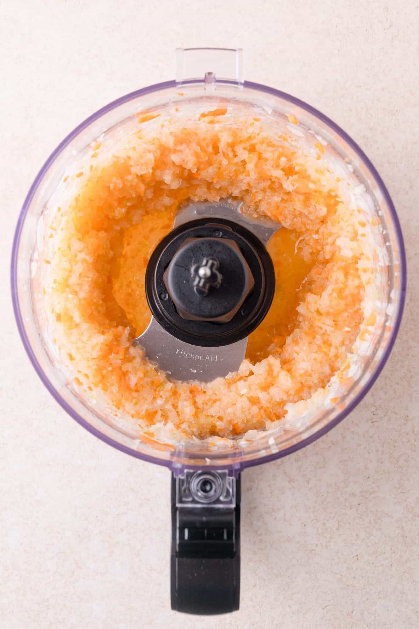 Overhead view of blended carrots, celery, and onions in a food processor on a light surface.