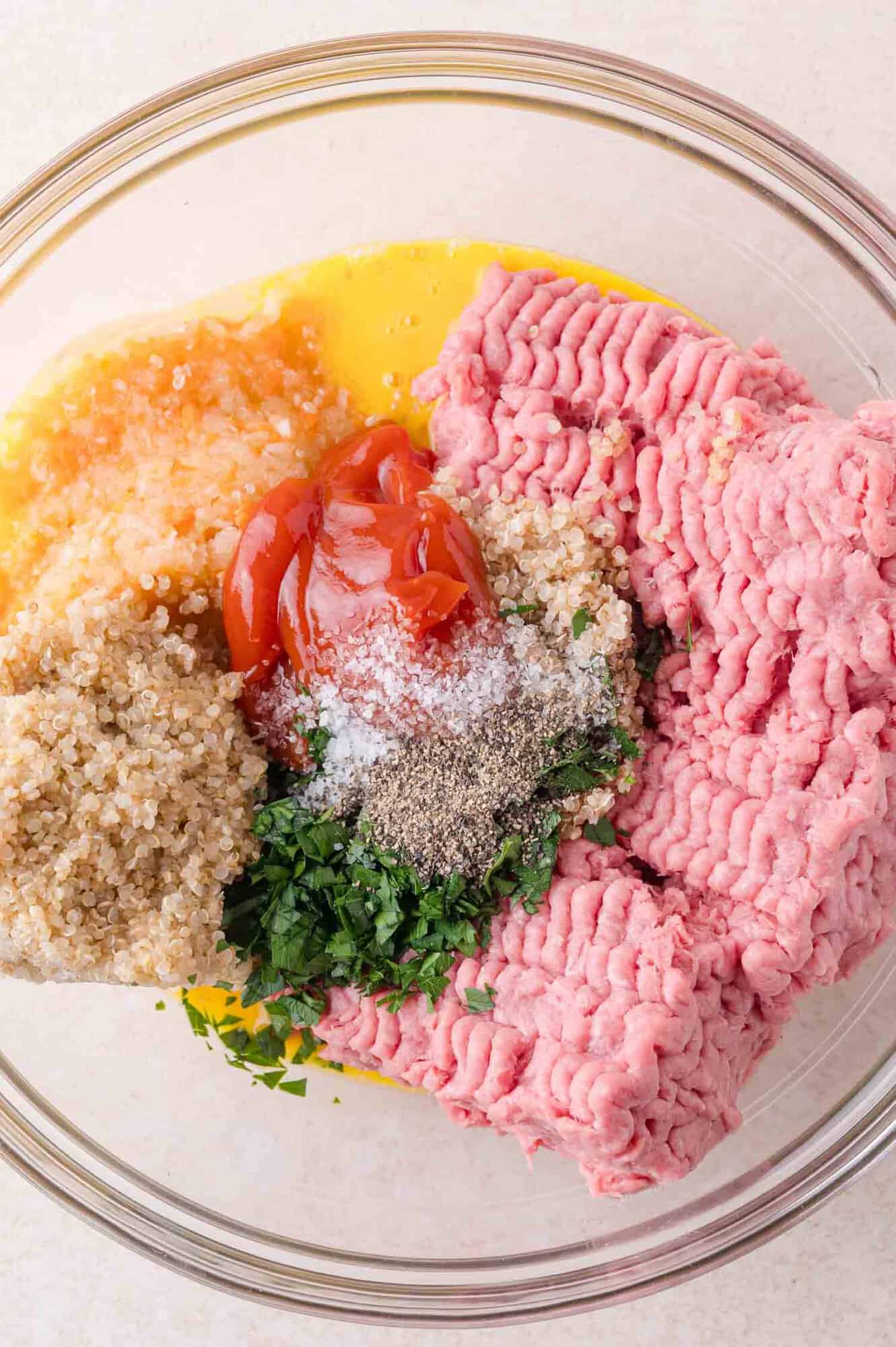 Ground turkey, egg, cooked quinoa, ketchup, chopped parsley, salt, and pepper in a clear mixing bowl on a light surface.
