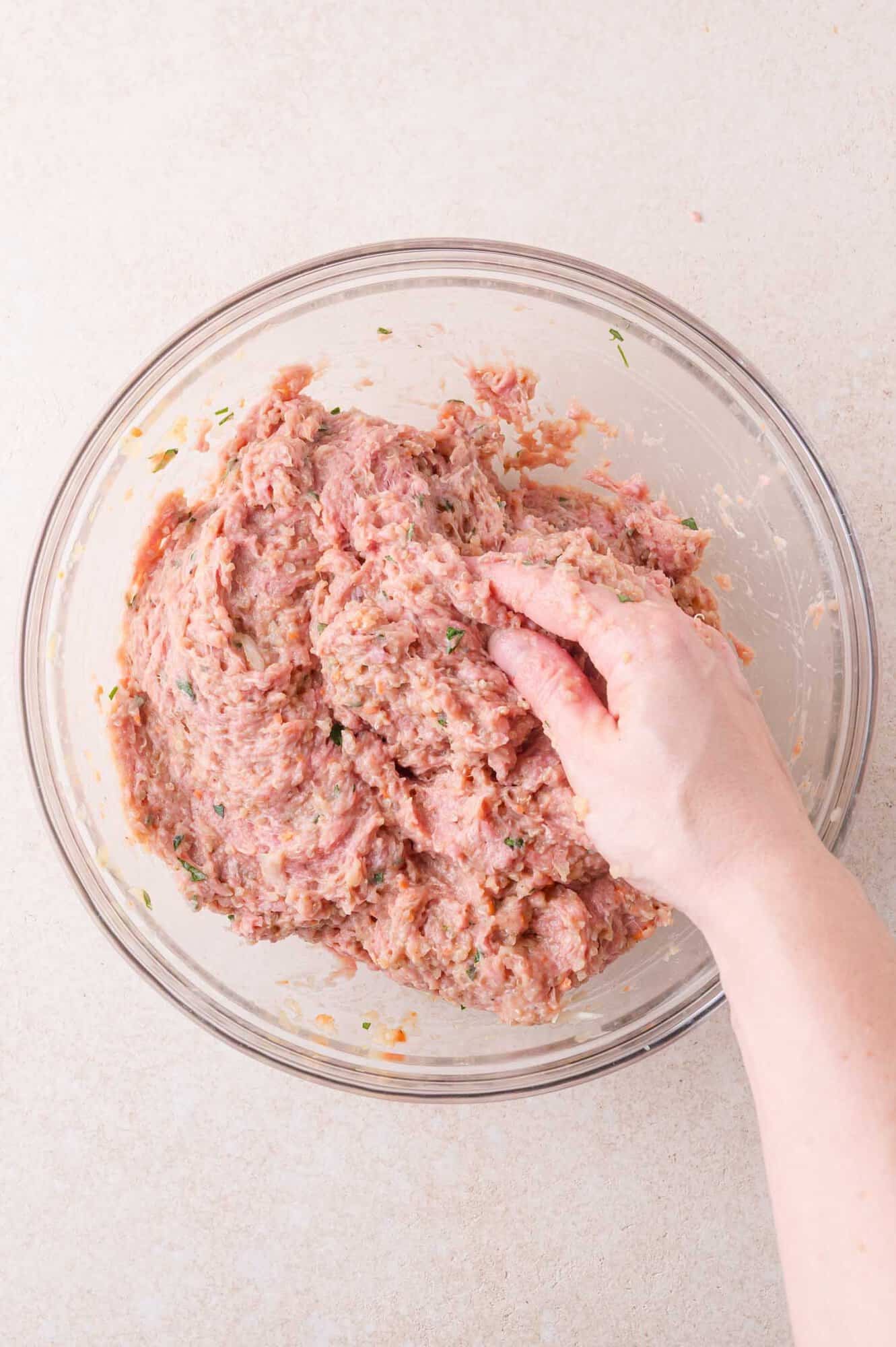 A hand mixes ground meat with herbs and seasoning in a clear glass bowl on a light countertop.