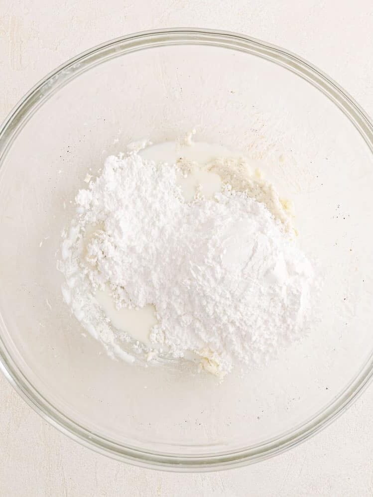 Powdered sugar added to a glass mixing bowl with butter, cream, and vanilla.