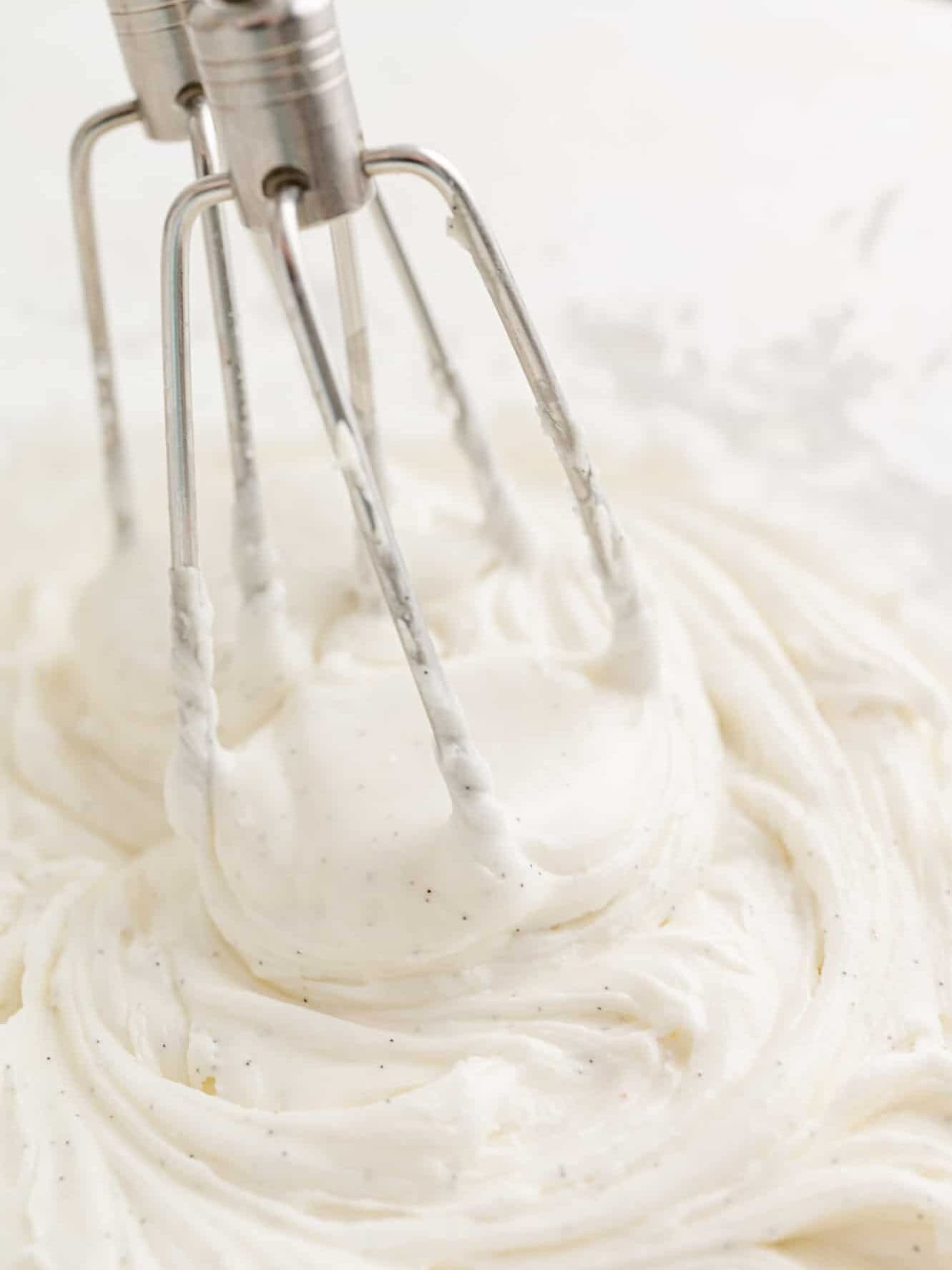 Two electric mixer attachments in a bowl of vanilla buttercream frosting.