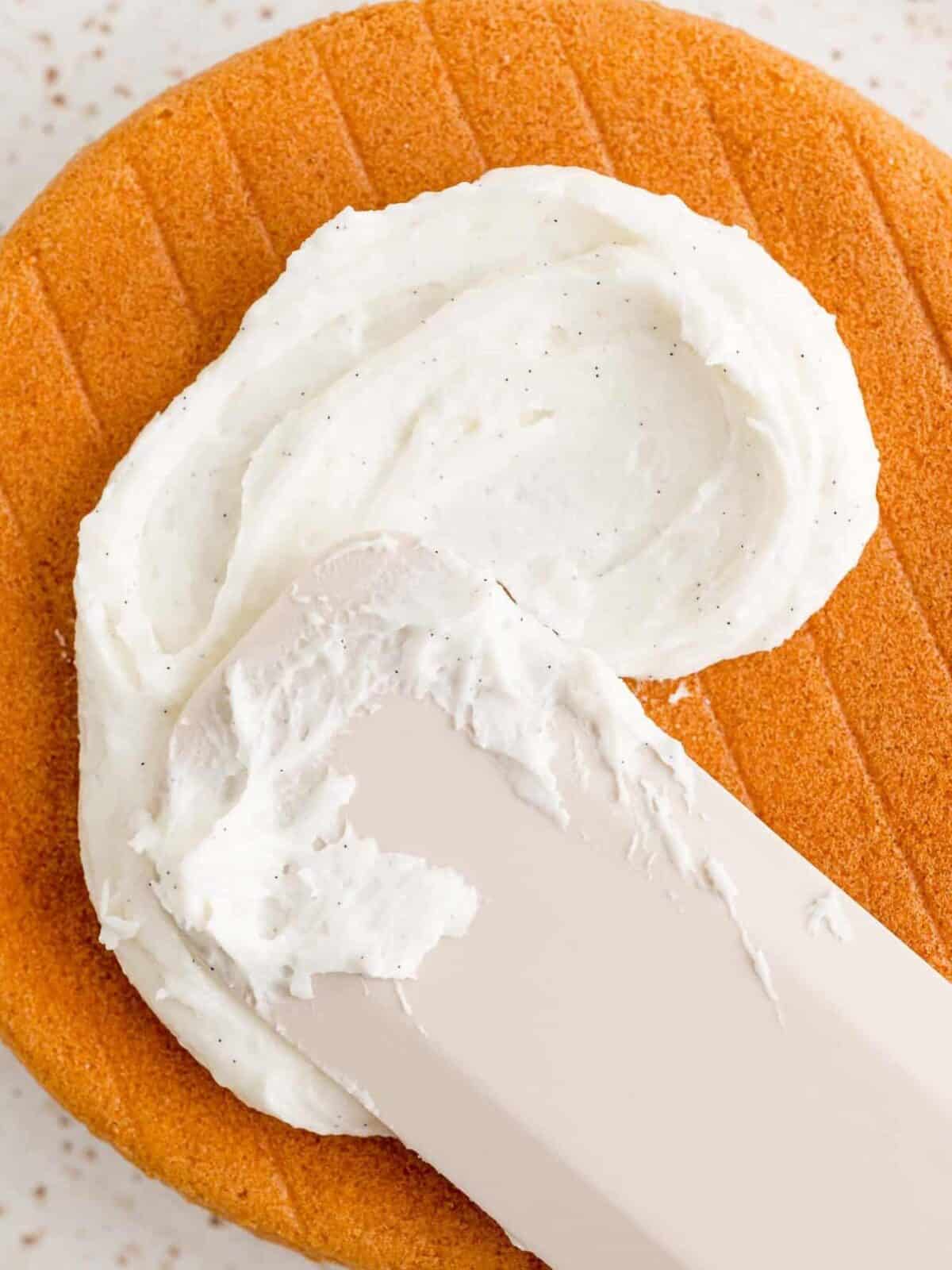 A spatula spreads vanilla buttercream over a vanilla cake layer.