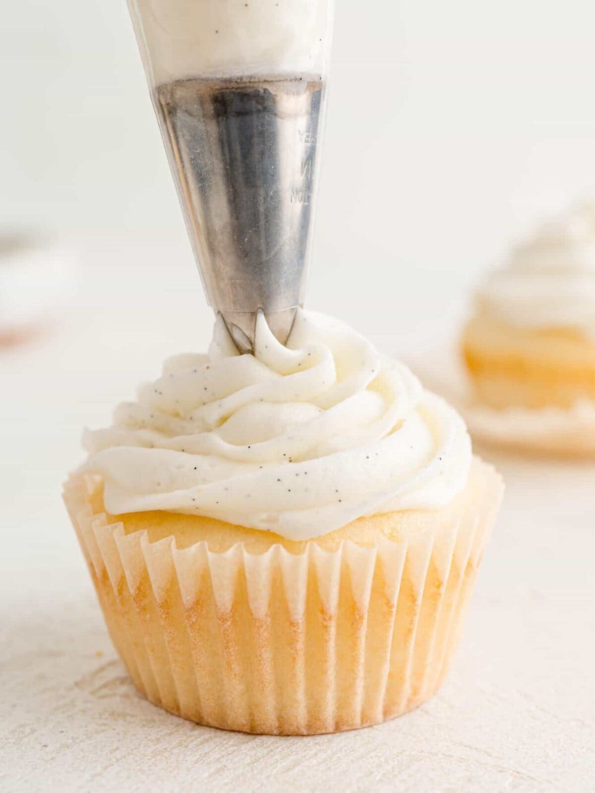 A piping tip pipes a swirl of vanilla buttercream onto a vanilla cupcake.