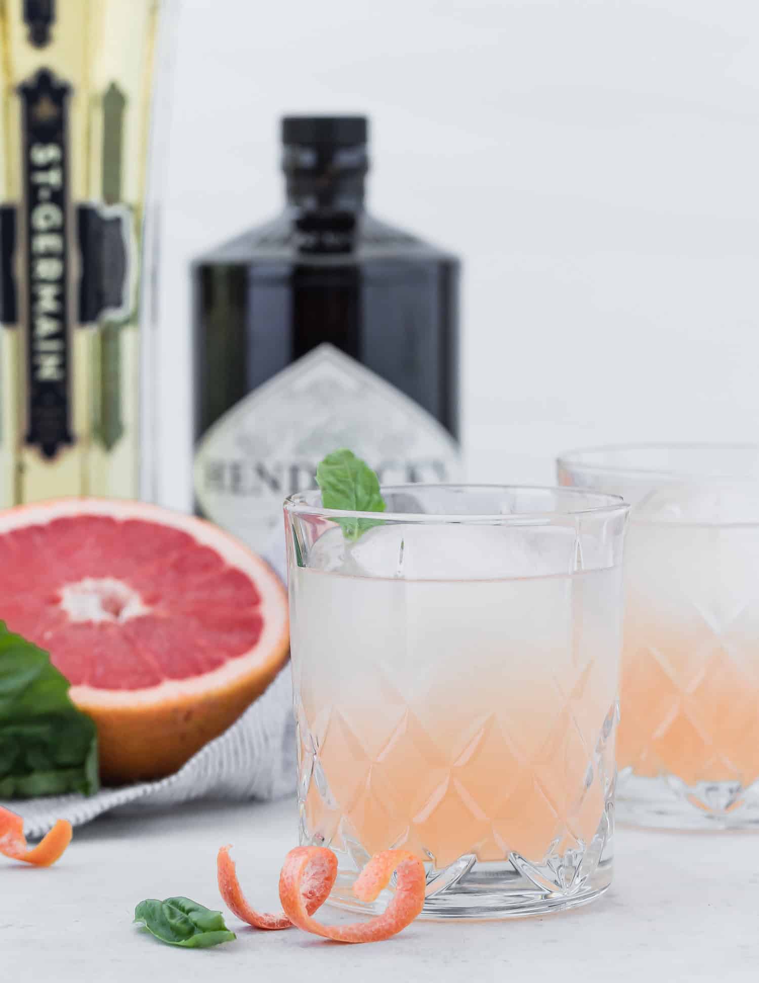 Two glasses of a light pink cocktail are on a white surface, garnished with mint and grapefruit peel, with a halved grapefruit and bottles of St-Germain and Hendricks gin in the background.