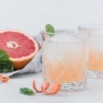 A glass of grapefruit cocktail with ice and a basil leaf garnish sits in front of a halved grapefruit, with grapefruit peel and basil leaves on a light surface.