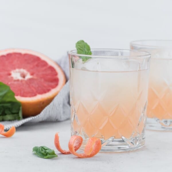 A glass of grapefruit cocktail with ice and a basil leaf garnish sits in front of a halved grapefruit, with grapefruit peel and basil leaves on a light surface.