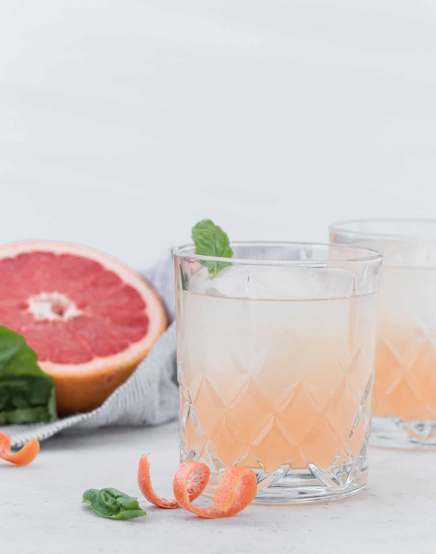 Crisp and refreshing, this gin, grapefruit, and elderflower cocktail is an easy one to make and an even easier one to drink! A glass of grapefruit cocktail garnished with basil, next to a grapefruit half, basil, and a grapefruit peel curl on a light surface.