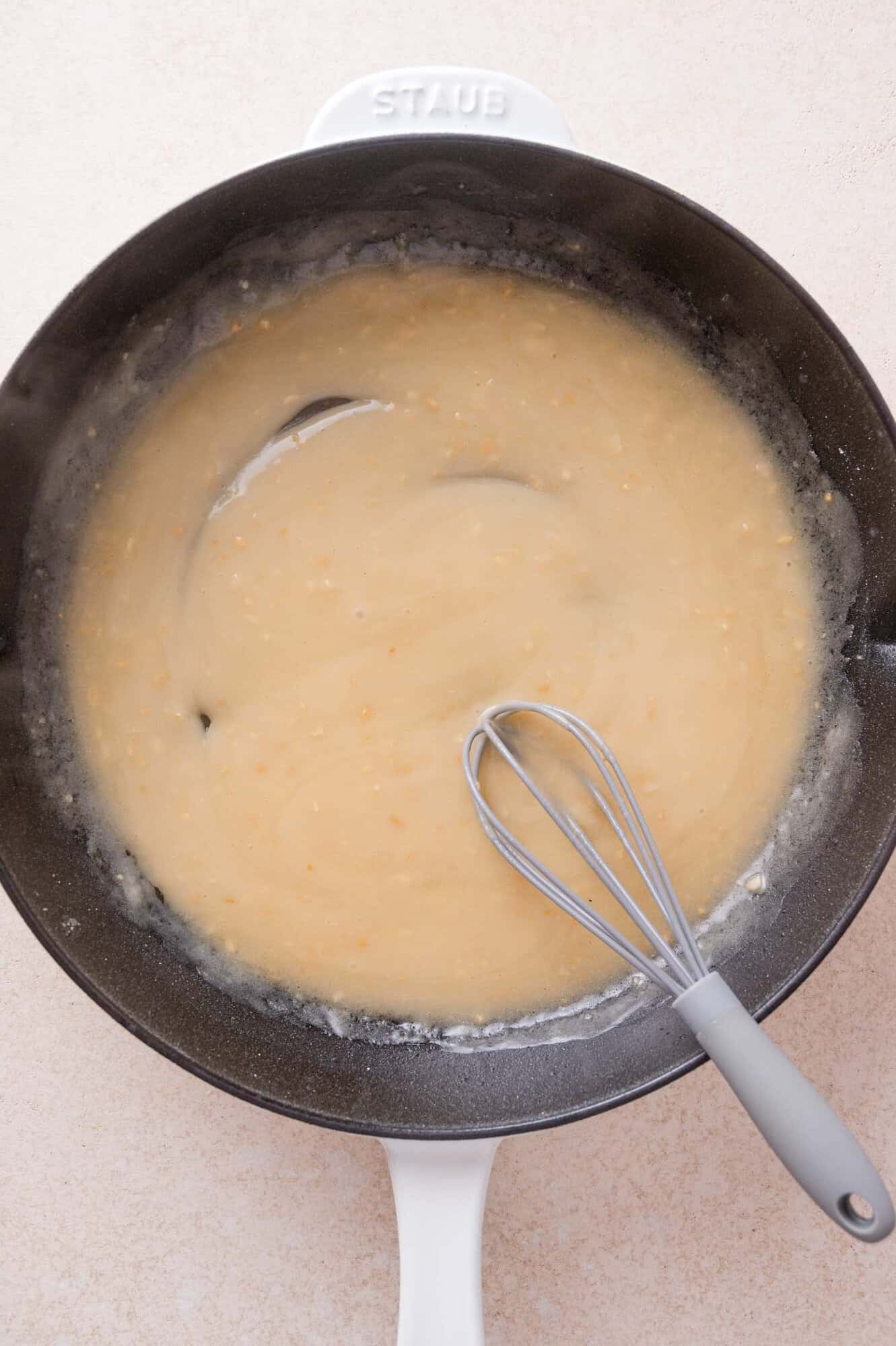 A whisk stirs a light brown sauce in a black skillet.