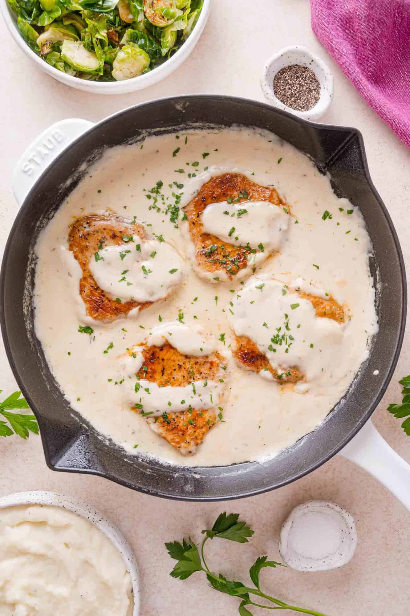 Four pork chops in a creamy white sauce inside a cast-iron skillet, served with a side of roasted Brussels sprouts and mashed potatoes.