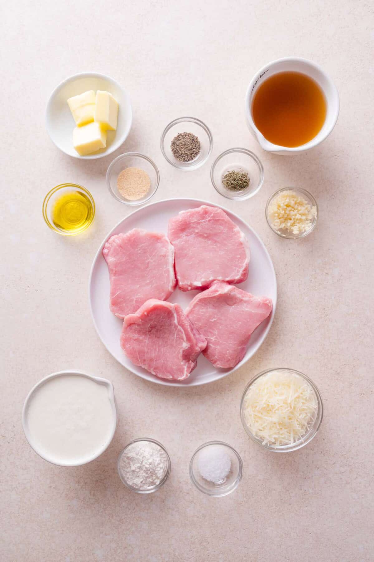 Four raw pork chops on a plate surrounded by bowls of butter, oil, cream, grated cheese, garlic, and various seasonings.