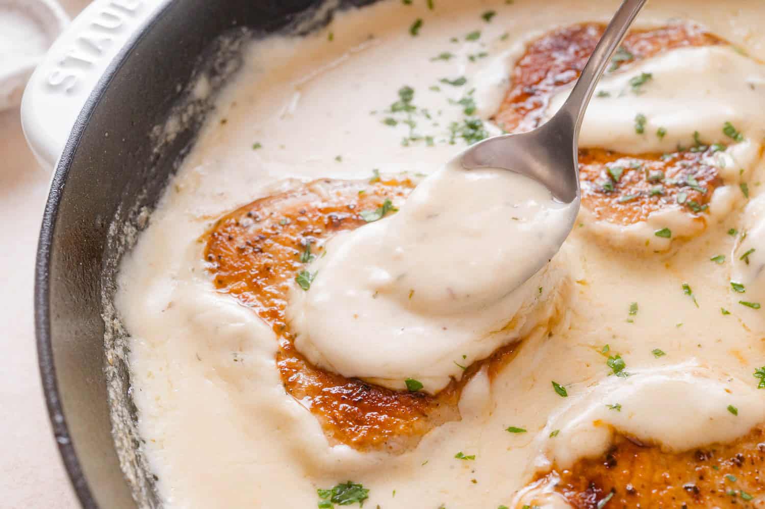 A spoon lifts creamy sauce from a skillet containing seared pork chops garnished with herbs.