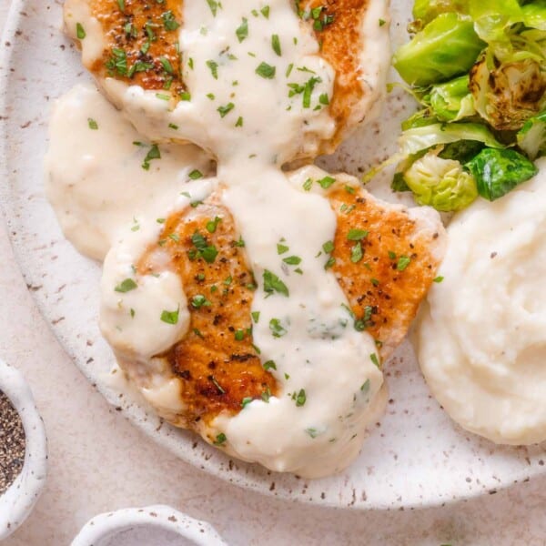 Two seared pork chops topped with creamy white sauce and herbs, served with mashed potatoes and roasted Brussels sprouts.