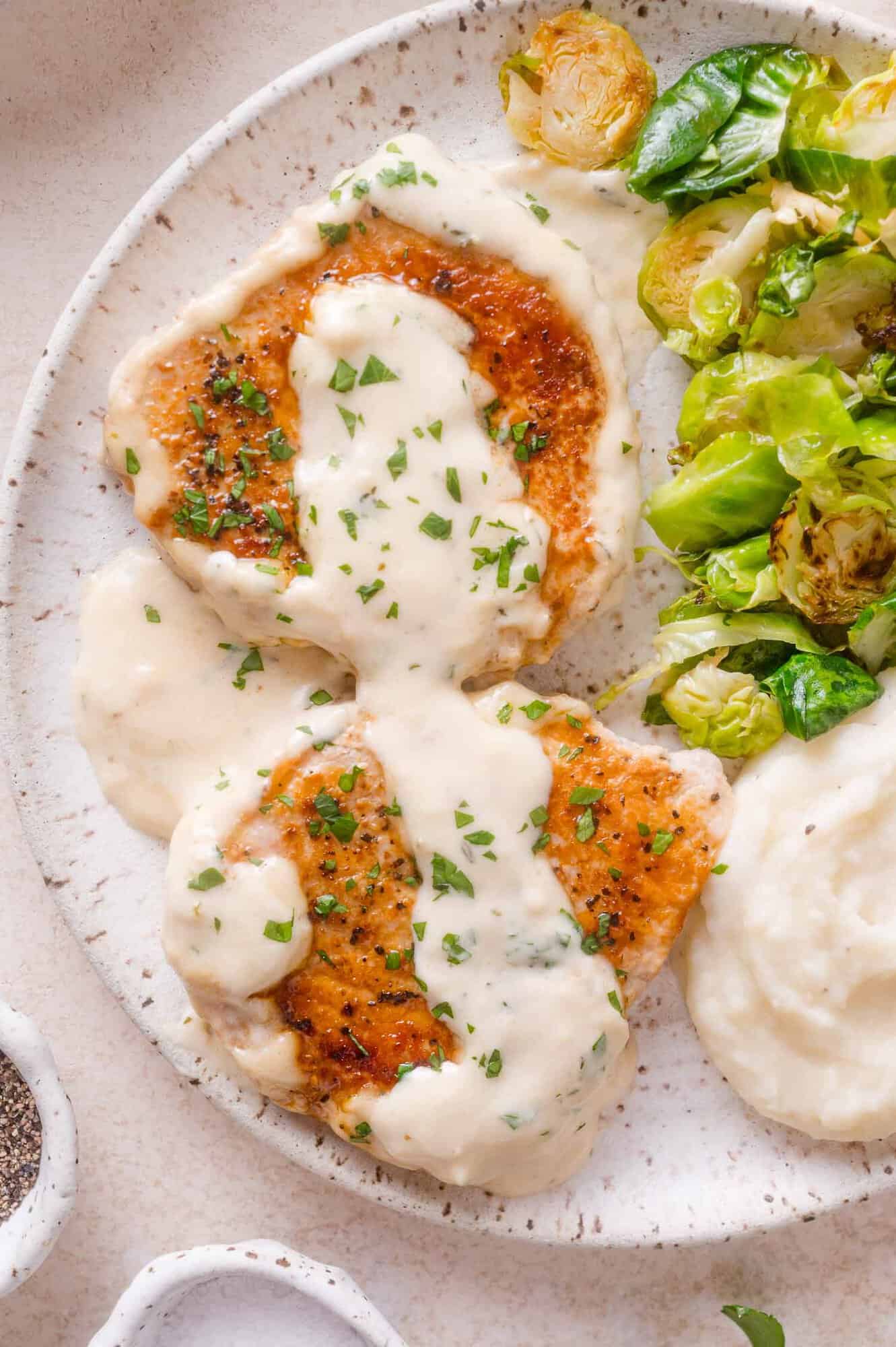 Two seared pork chops topped with creamy white sauce and herbs, served with roasted Brussels sprouts and mashed potatoes on a speckled plate.