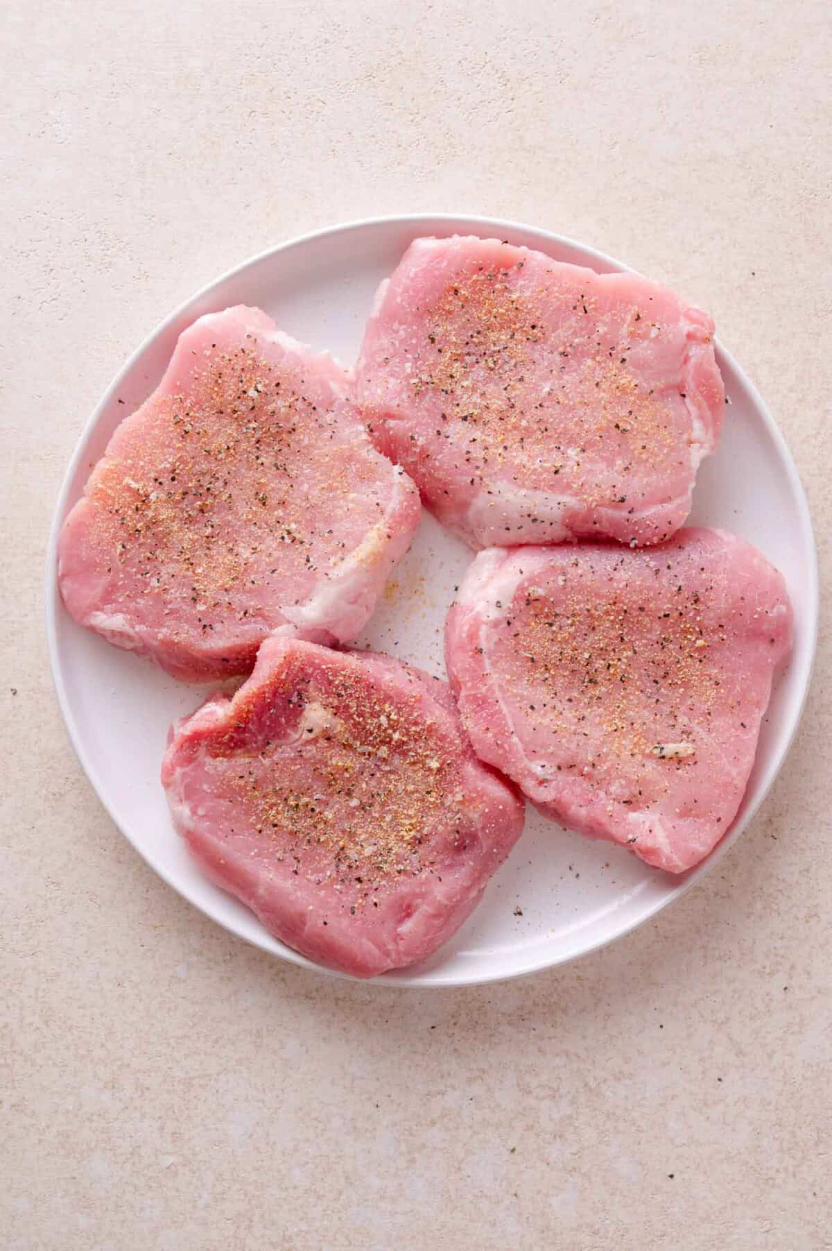 Four raw pork chops seasoned with salt and pepper on a white plate.