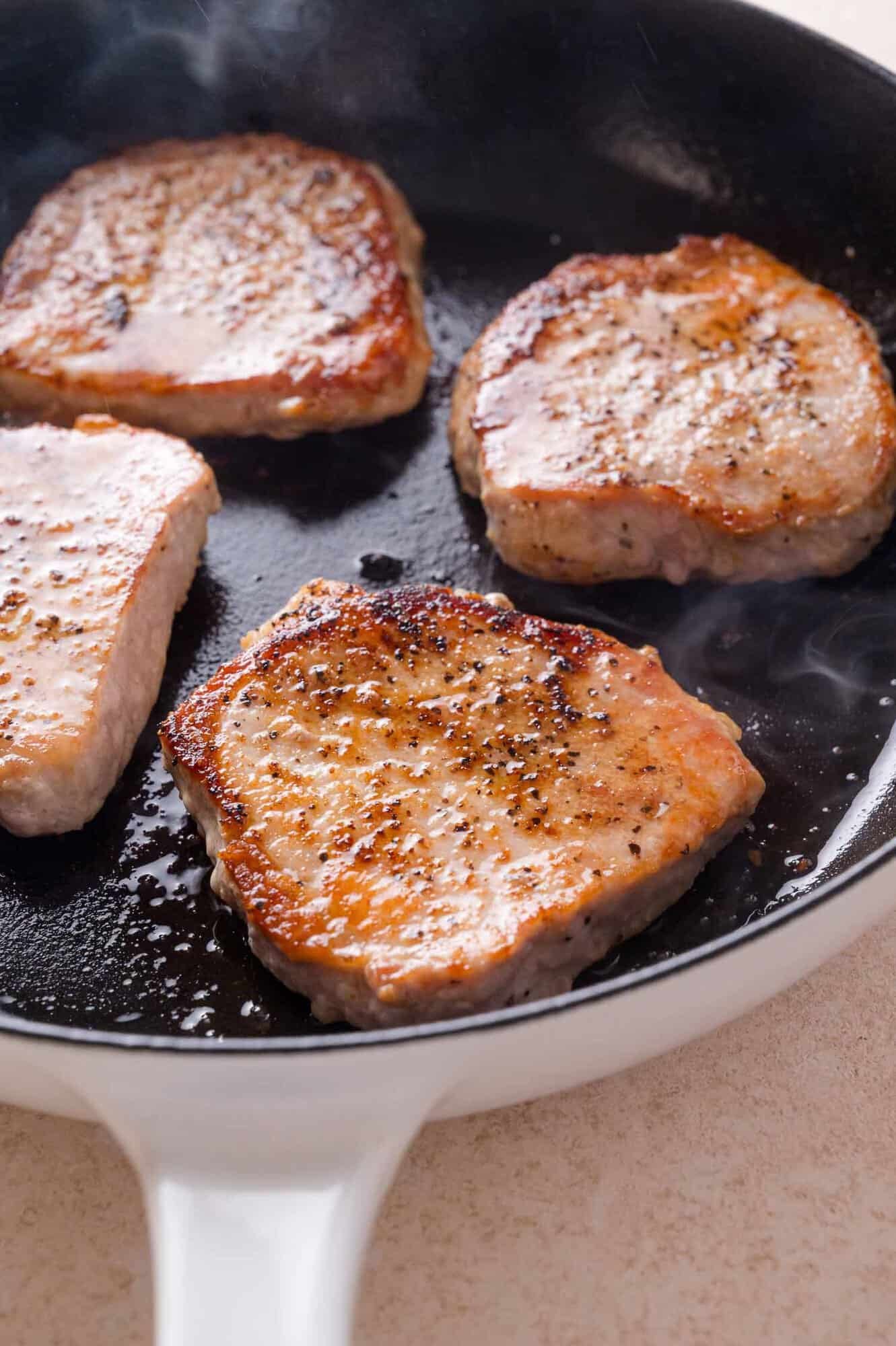 Four seasoned pork chops searing in a skillet.