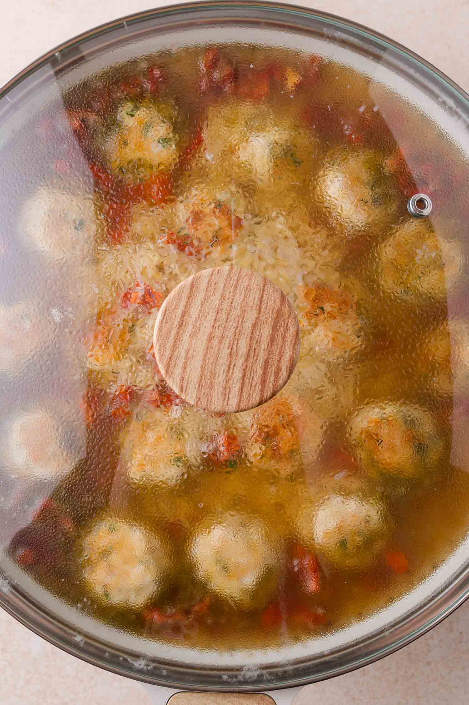 A glass lid covering a pot of orzo containing meatballs and chopped vegetables.