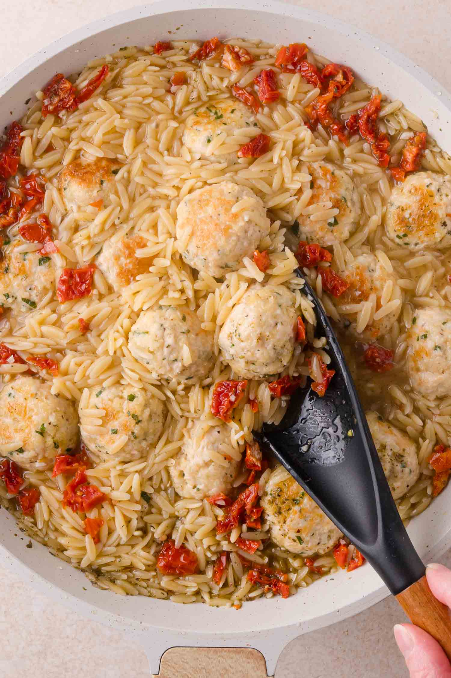 A pan of orzo pasta with meatballs and sun-dried tomatoes being stirred by a spatula.