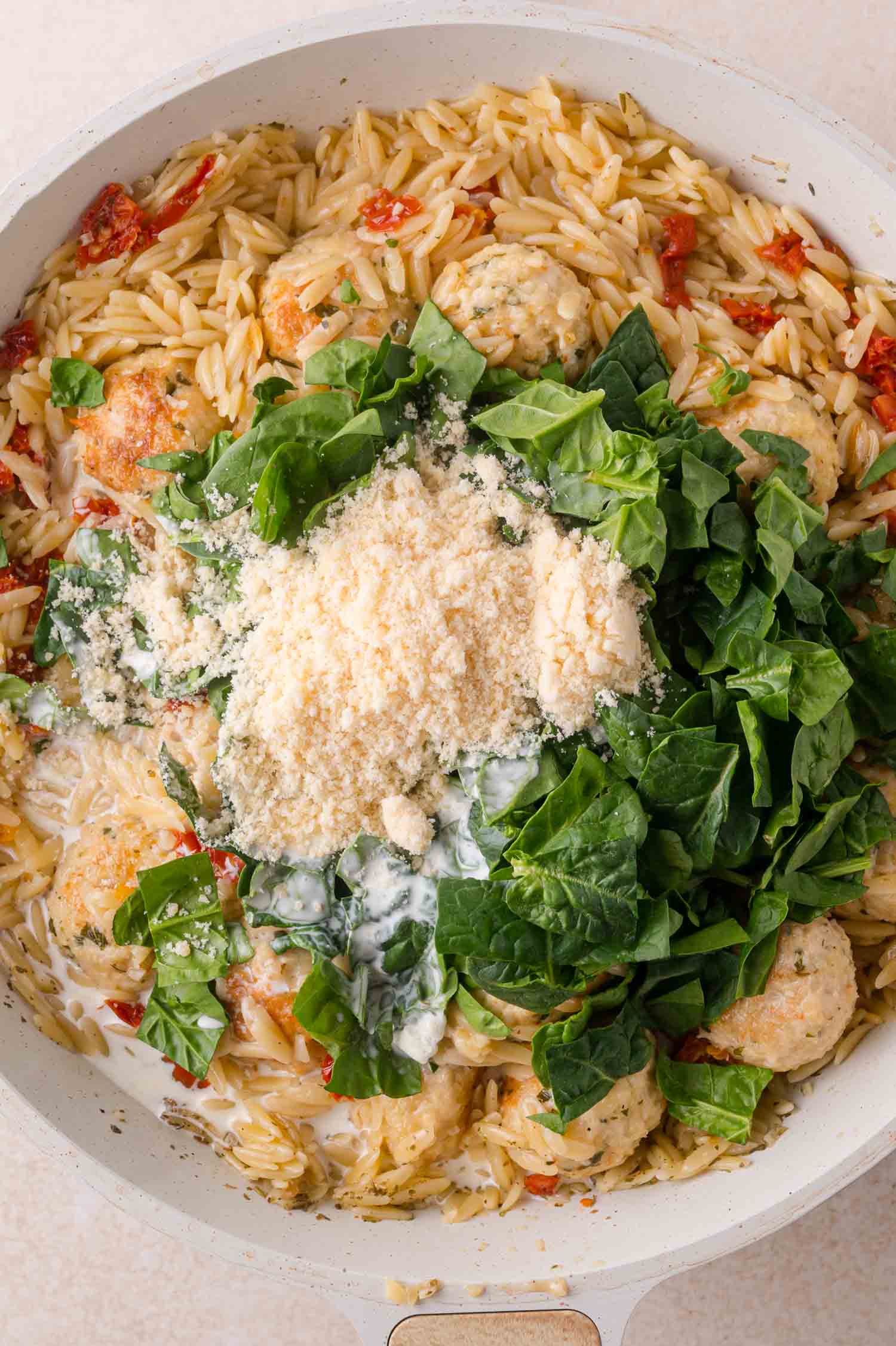 Orzo pasta with meatballs, sun-dried tomatoes, spinach, and a mound of grated cheese in a skillet.