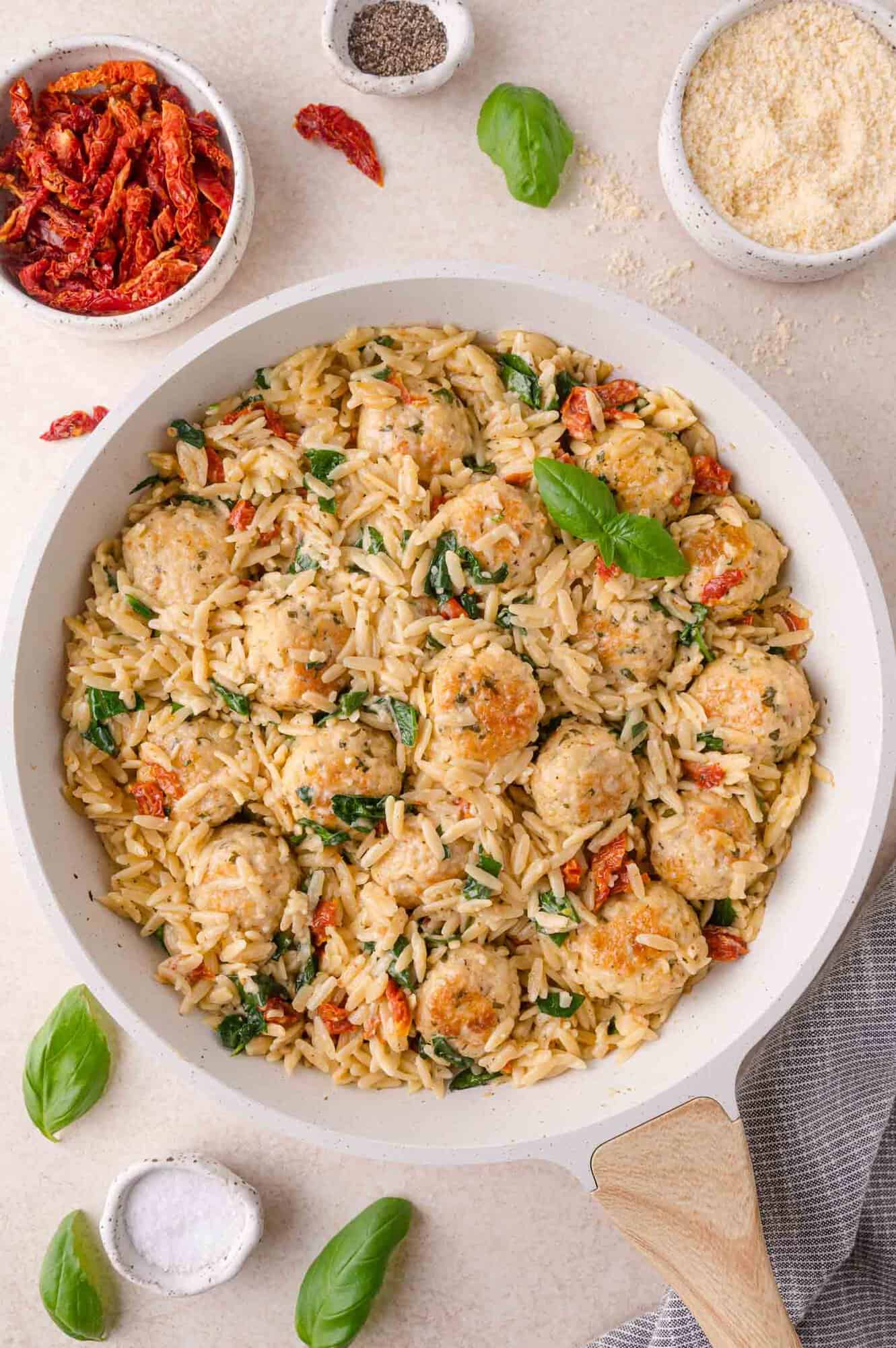 A skillet of orzo pasta with meatballs, spinach, and sun-dried tomatoes, surrounded by bowls of parmesan, sun-dried tomatoes, and basil leaves.