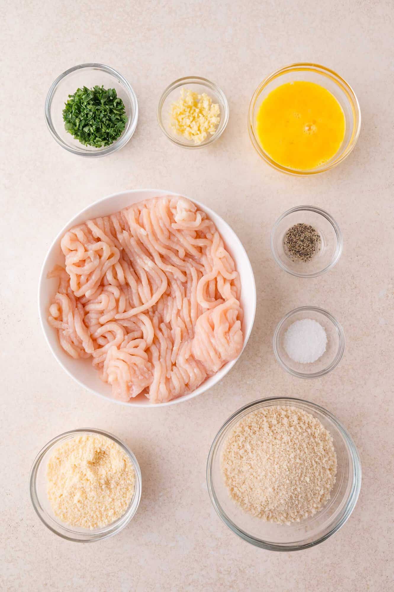 A large bowl of ground chicken surrounded by small bowls containing chopped parsley, minced garlic, beaten egg, black pepper, salt, grated cheese, and breadcrumbs.