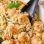 A black spoon lifting a portion of orzo pasta tossed with chicken meatballs, spinach, and sun-dried tomatoes.