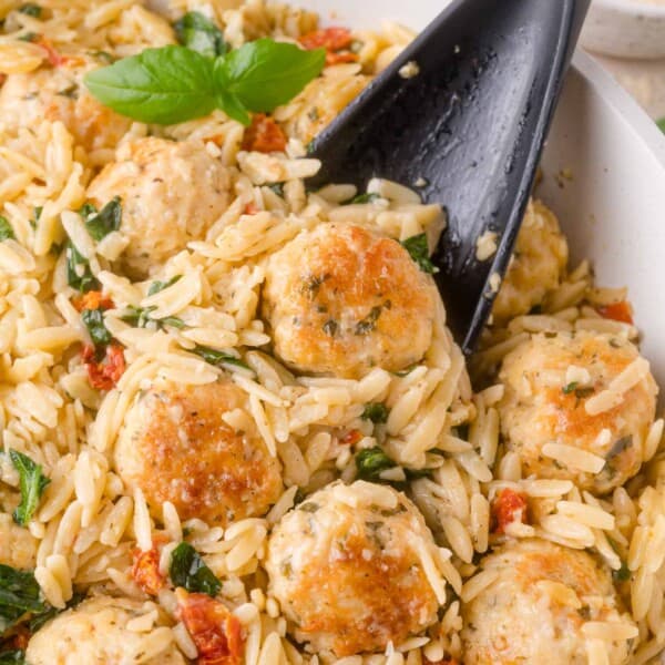 A black spoon lifting a portion of orzo pasta tossed with chicken meatballs, spinach, and sun-dried tomatoes.