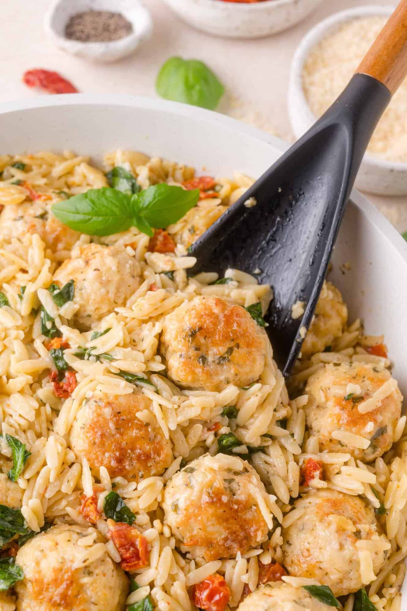 A black spoon lifts meatballs and orzo with spinach and sun-dried tomatoes from a white pan.