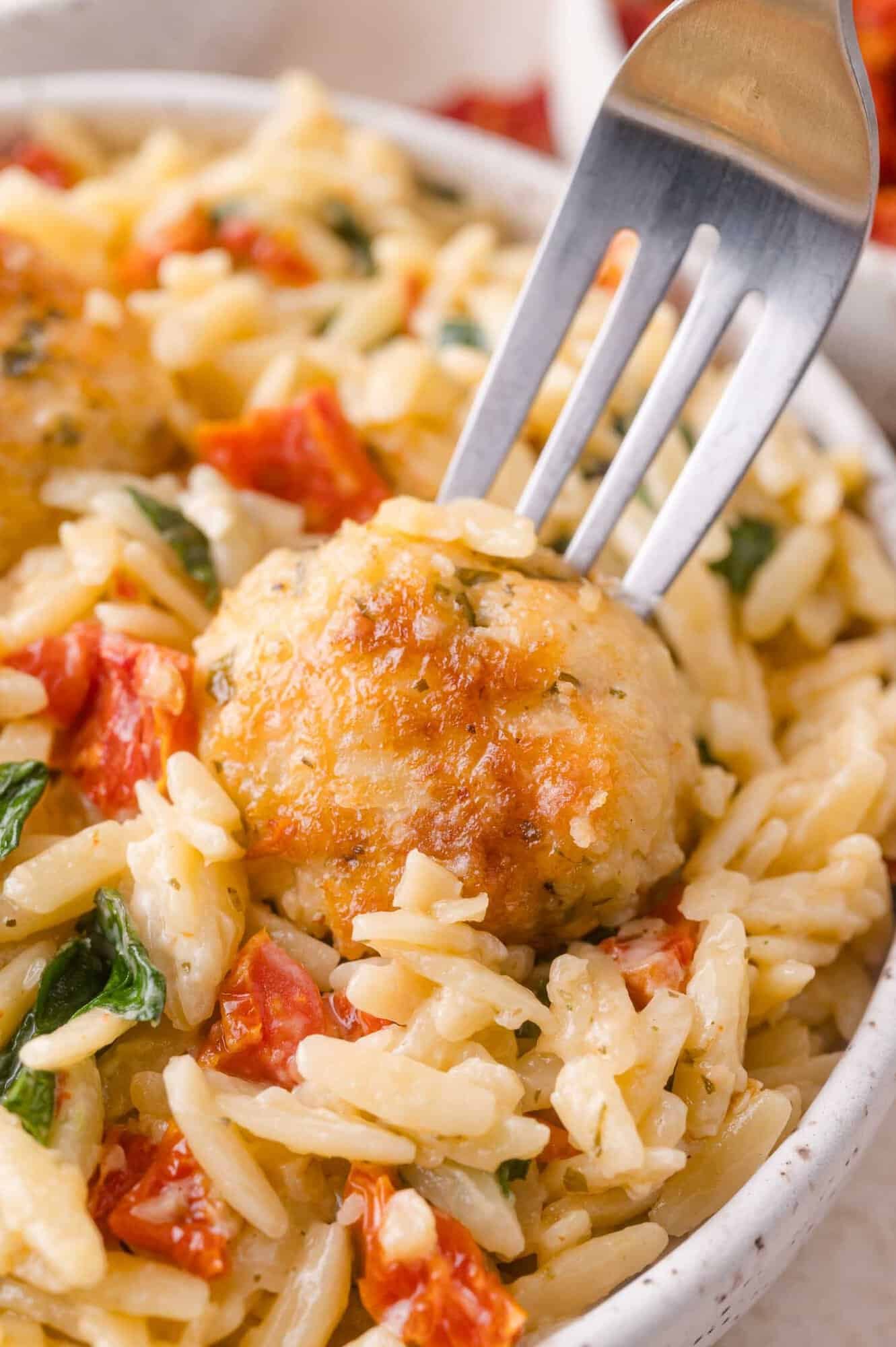 A fork lifting a golden-brown meatball from a bowl of orzo pasta with tomatoes and spinach.