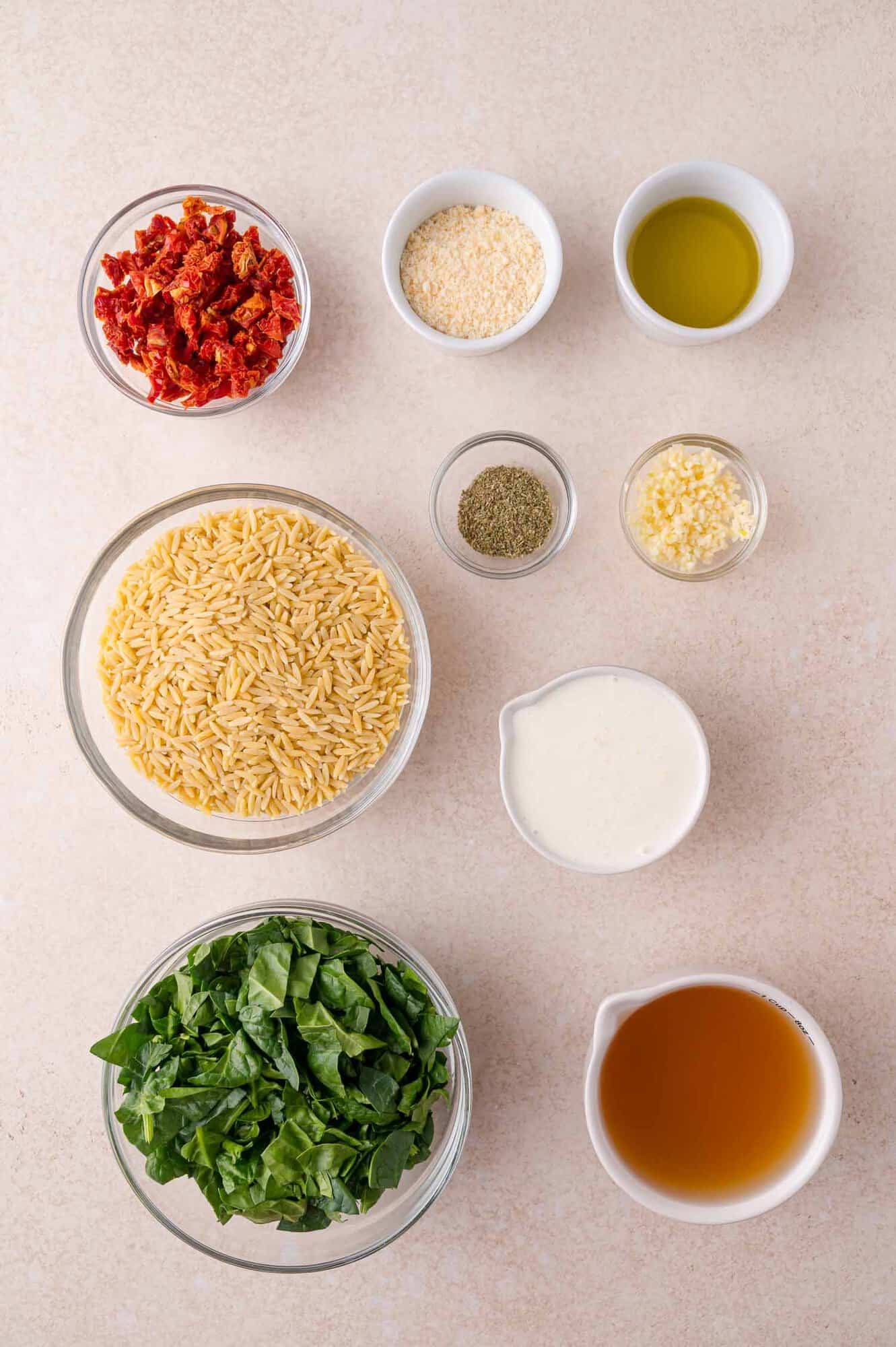 Ingredients for a pasta dish arranged on a light surface: sun-dried tomatoes, grated cheese, olive oil, orzo, herbs, minced garlic, cream, spinach, and broth.