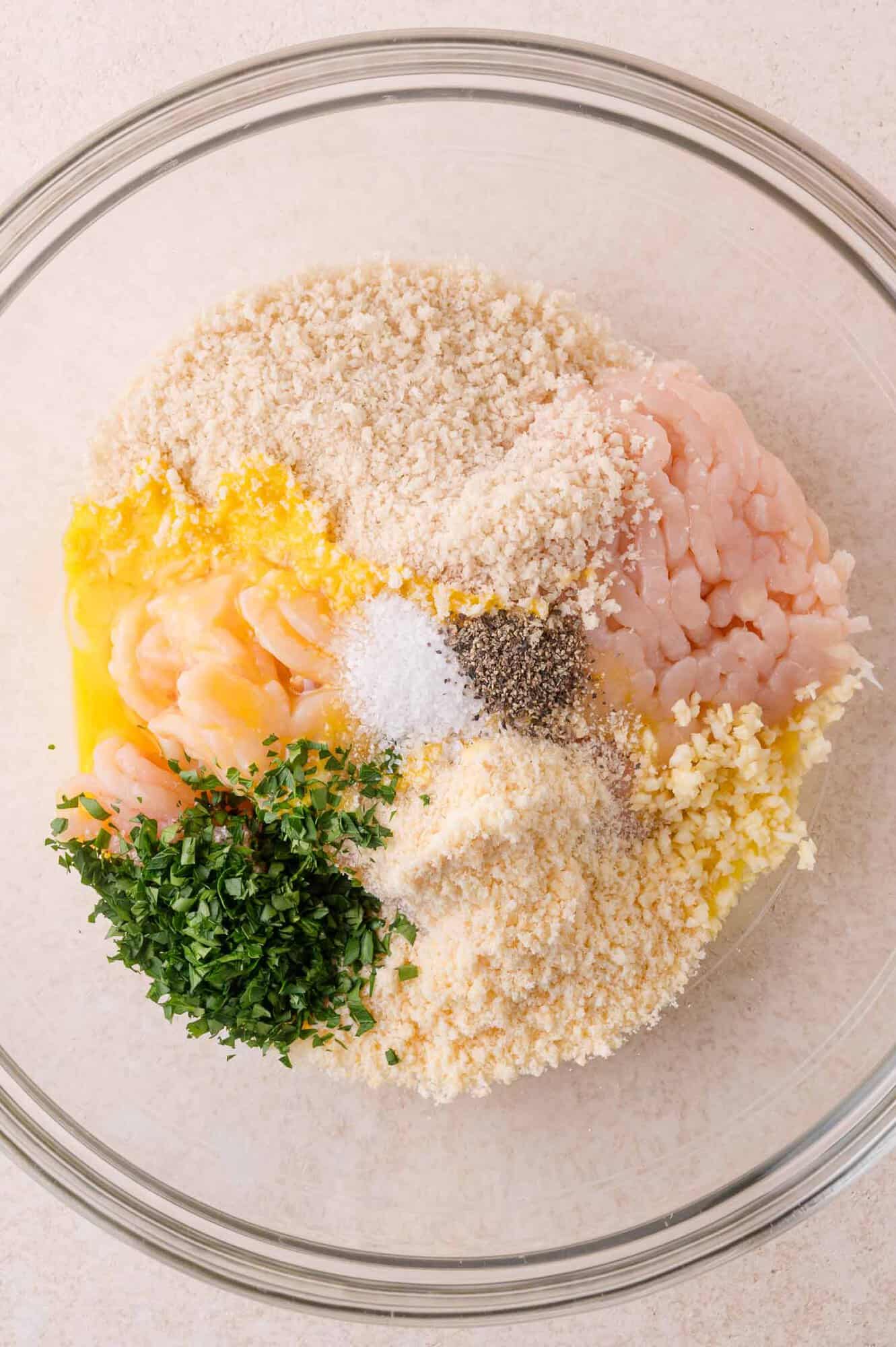 A glass bowl containing raw ground chicken, breadcrumbs, egg, minced garlic, fresh parsley, salt, and pepper.