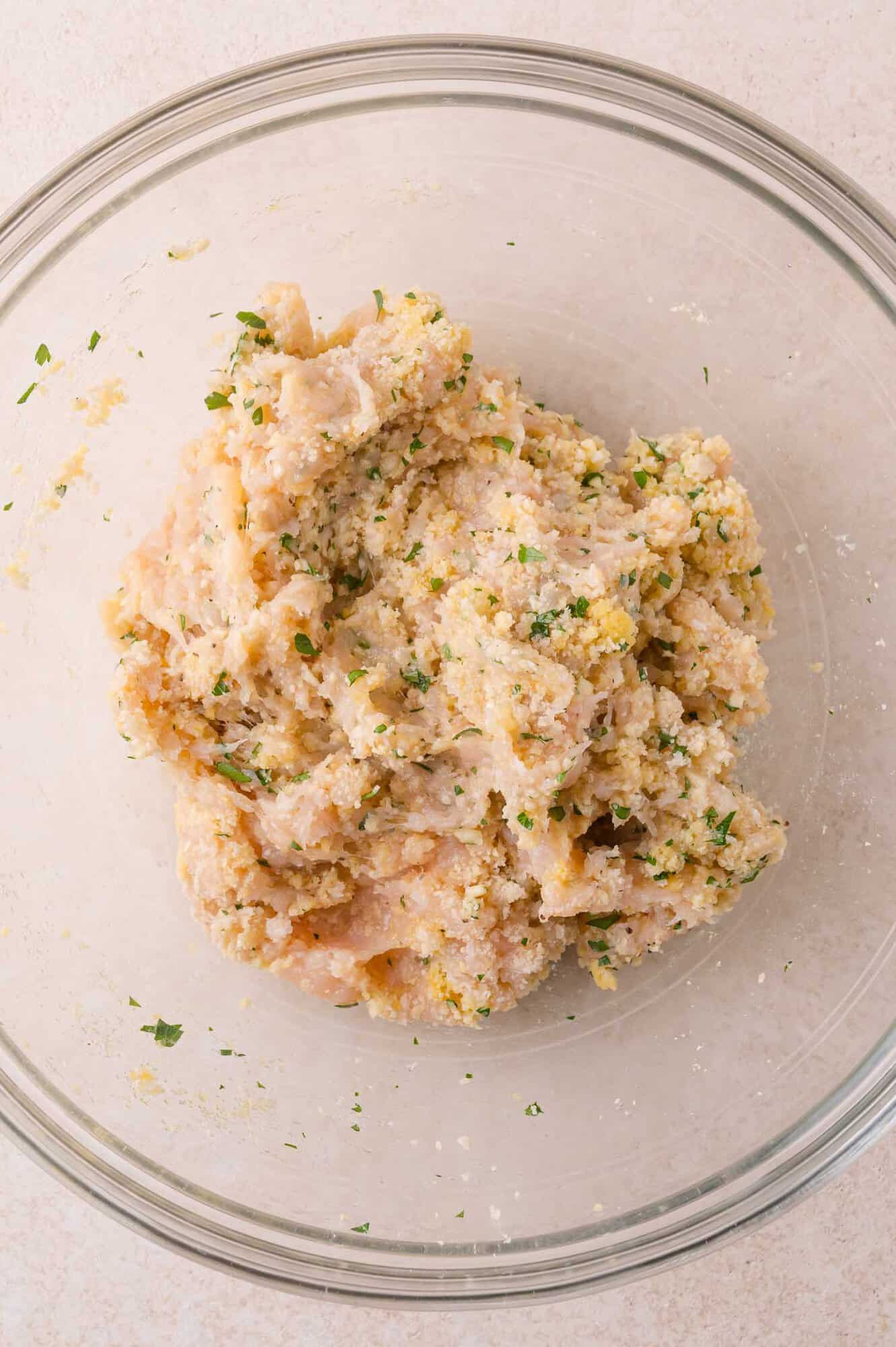Raw ground chicken mixed with breadcrumbs and herbs in a glass bowl.