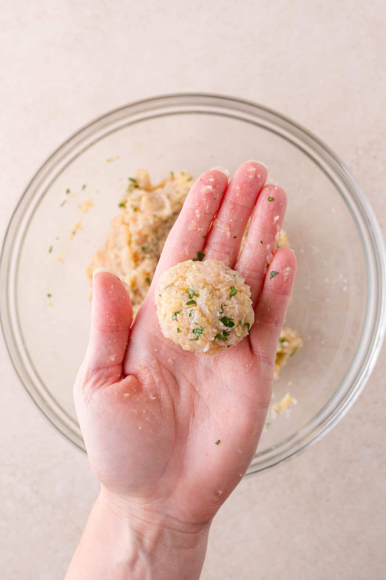 A hand holding a raw, meatball over a glass mixing bowl.