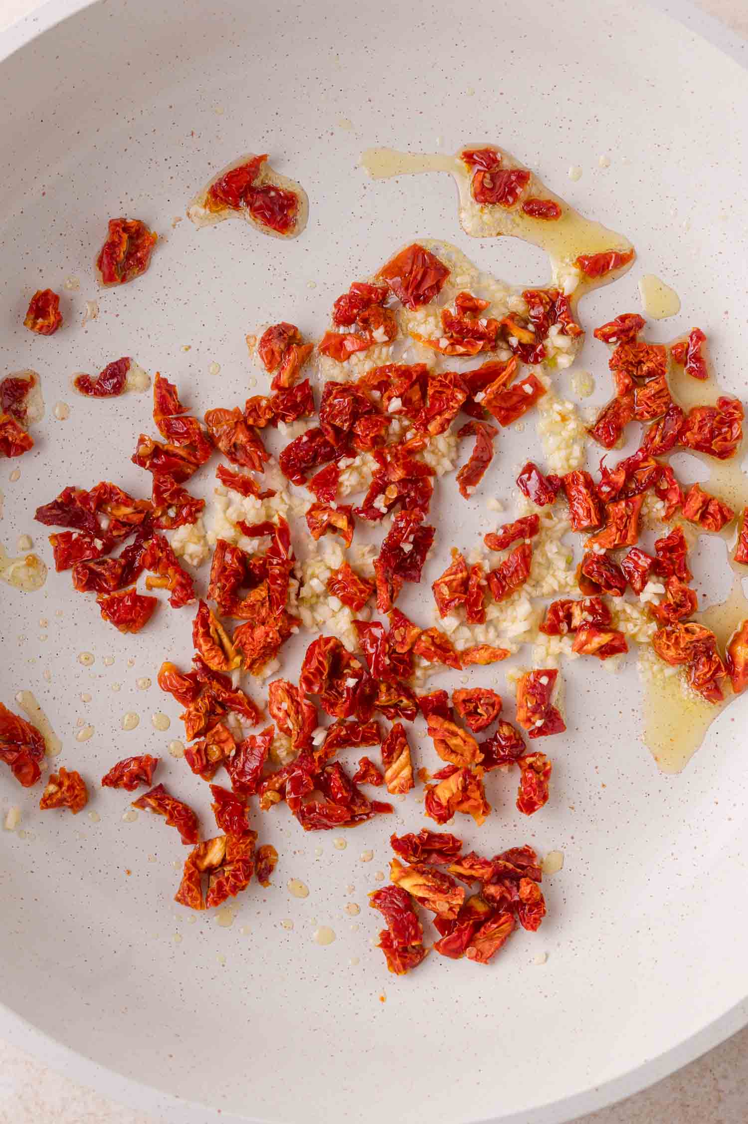 Chopped sun-dried tomatoes and minced garlic sautéing in oil in a white skillet.