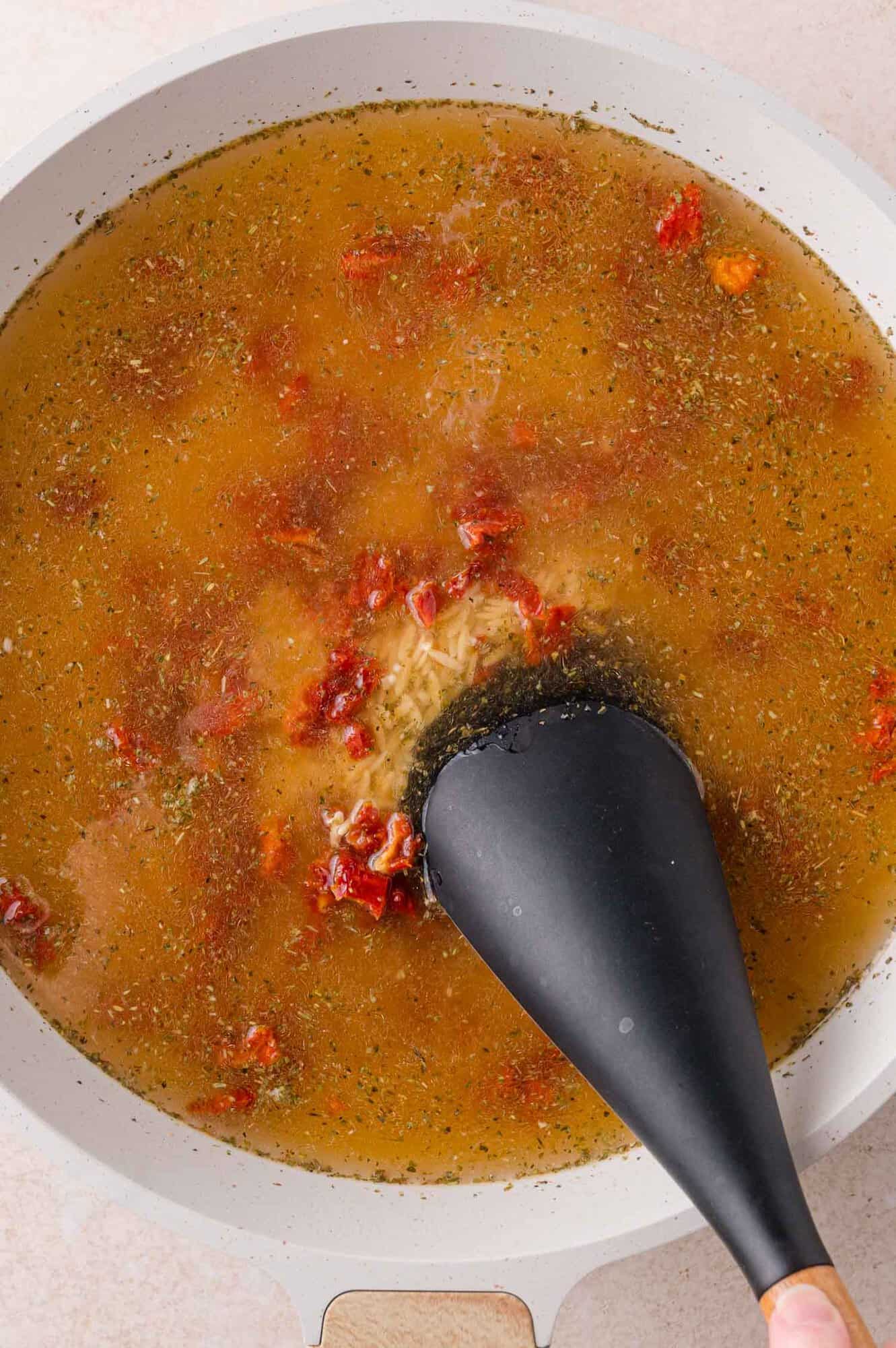 A black spoon stirs orzo, broth, and sun-dried tomatoes in a wide skillet.