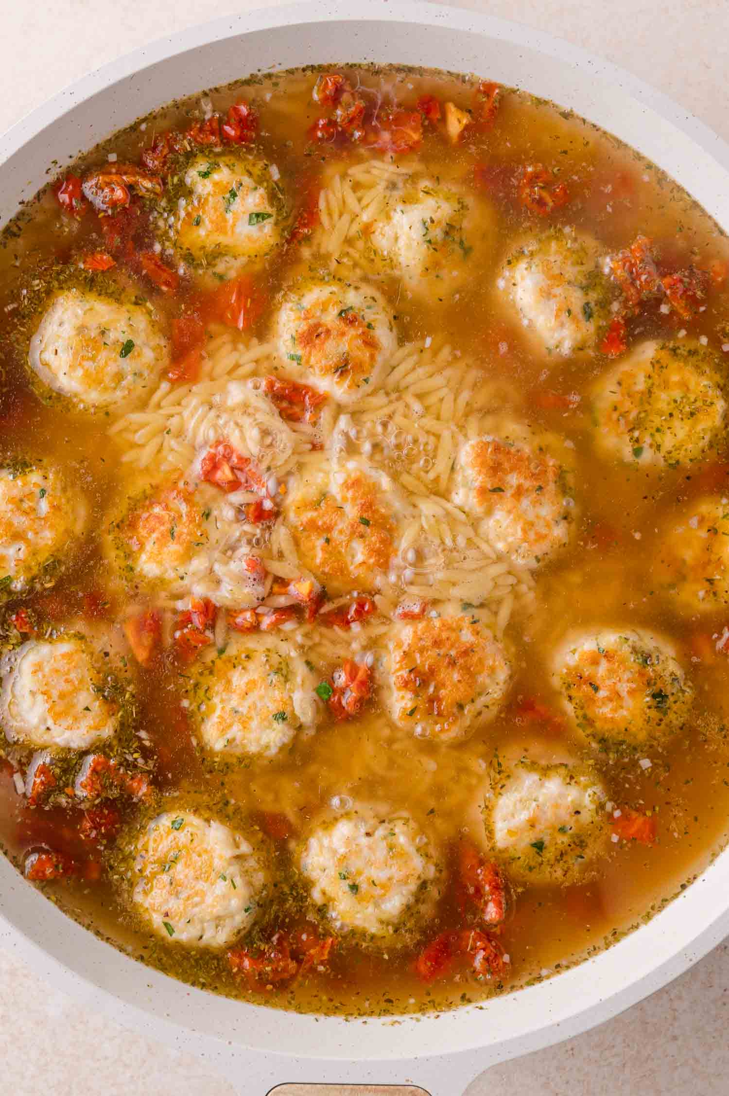 Meatballs and orzo pasta simmering in a clear broth with bits of sun-dried tomato and herbs.
