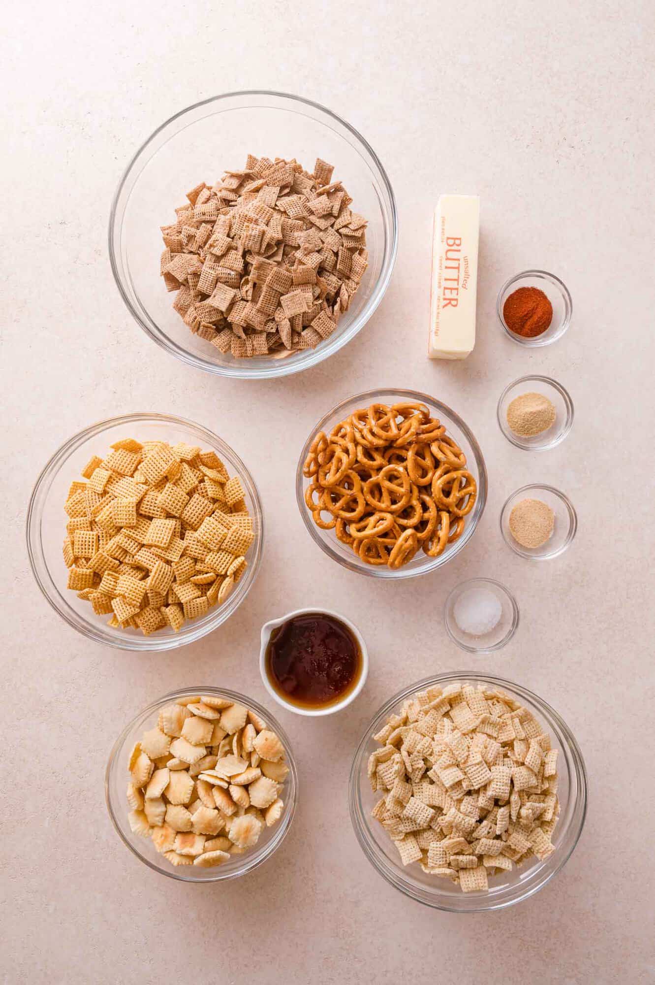 Ingredients for Chex mix arranged in bowls, including three types of cereal, pretzels, crackers, a stick of butter, and small bowls of spices and salt.