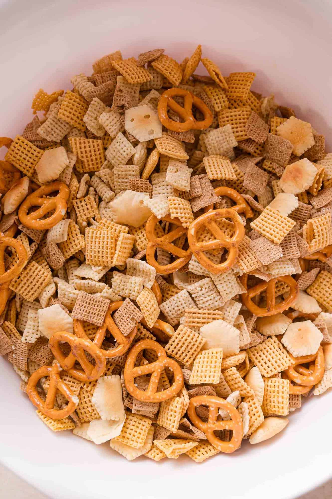 A bowl of snack mix ingredients containing various types of cereal squares, pretzels, and small crackers.