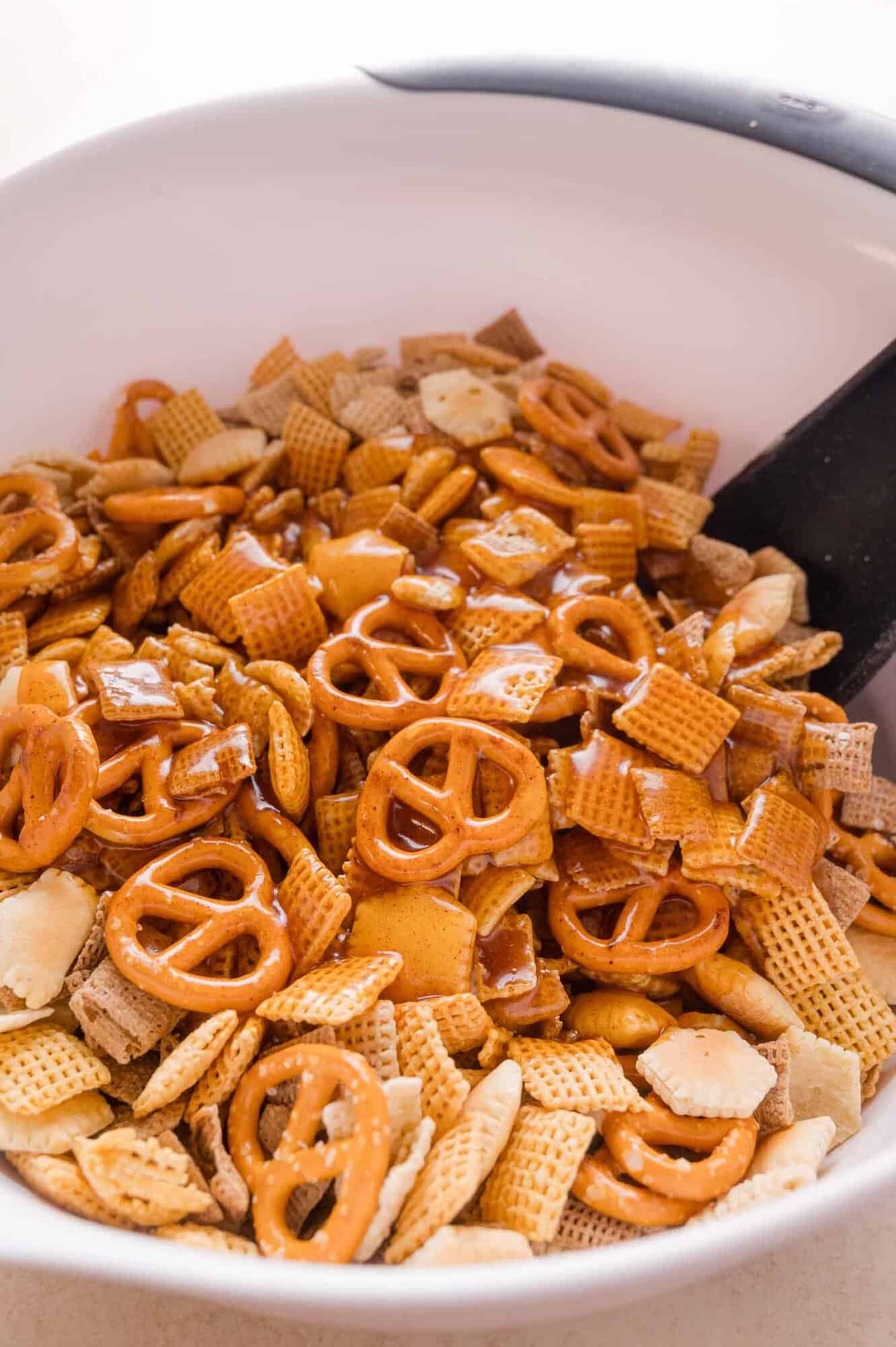 A mixture of cereal squares, pretzels, and crackers coated in melted butter and spices in a white bowl with a spatula.
